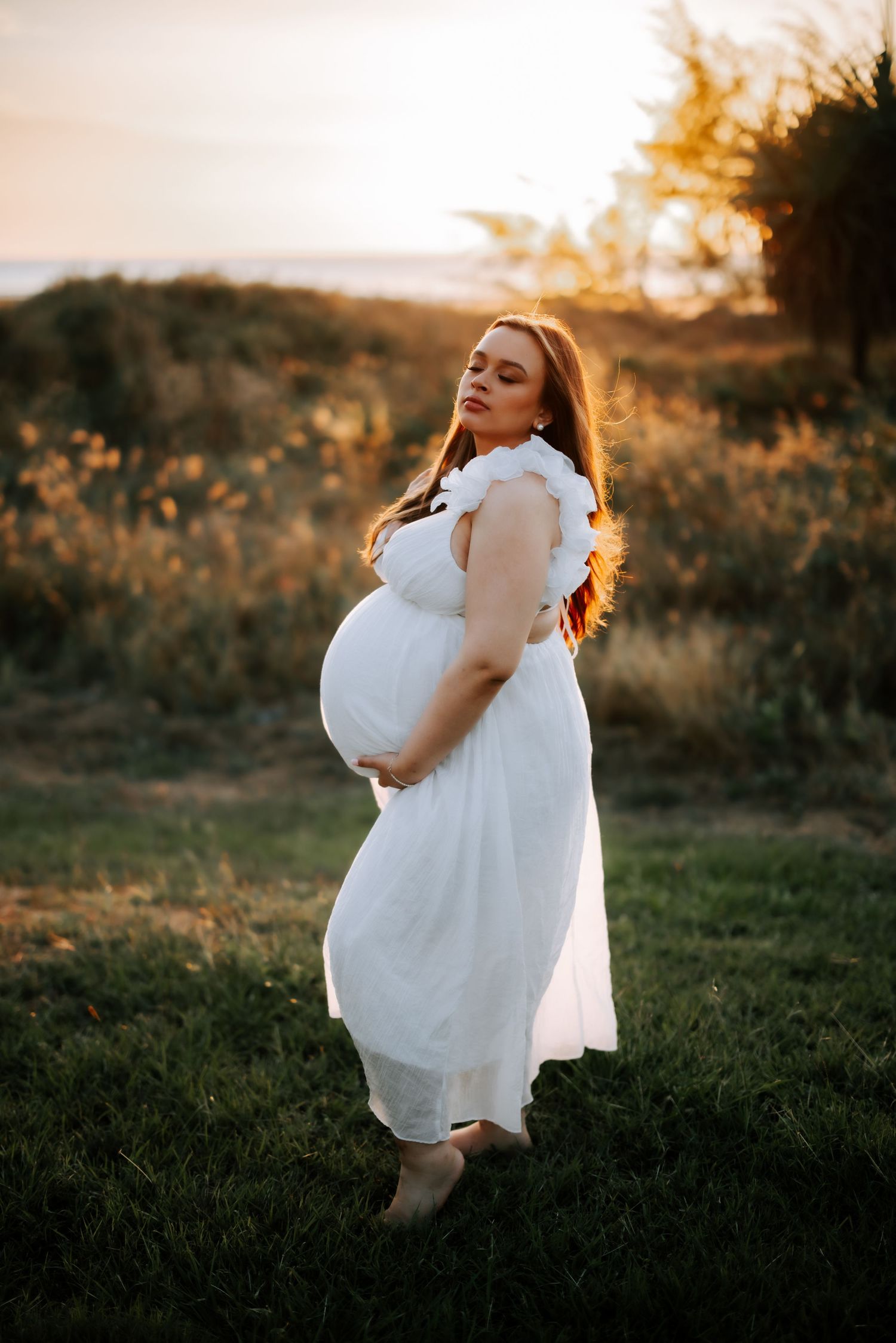 Pregnant woman in white dress holding baby bump in golden sunset field outdoors.