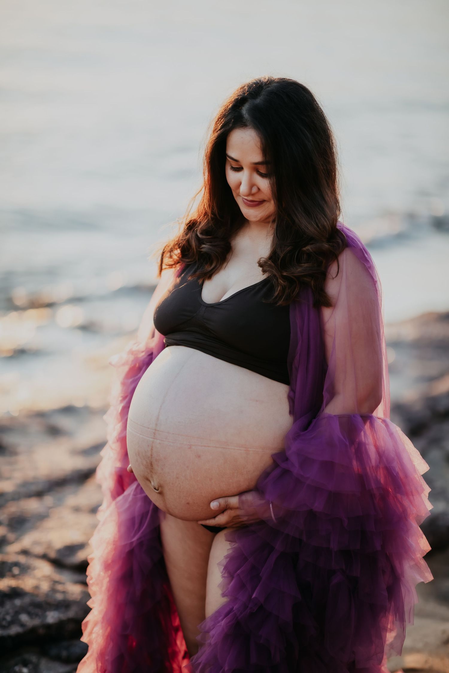 Pregnant woman in purple tulle robe tenderly cradling bare baby bump on rocky beach.