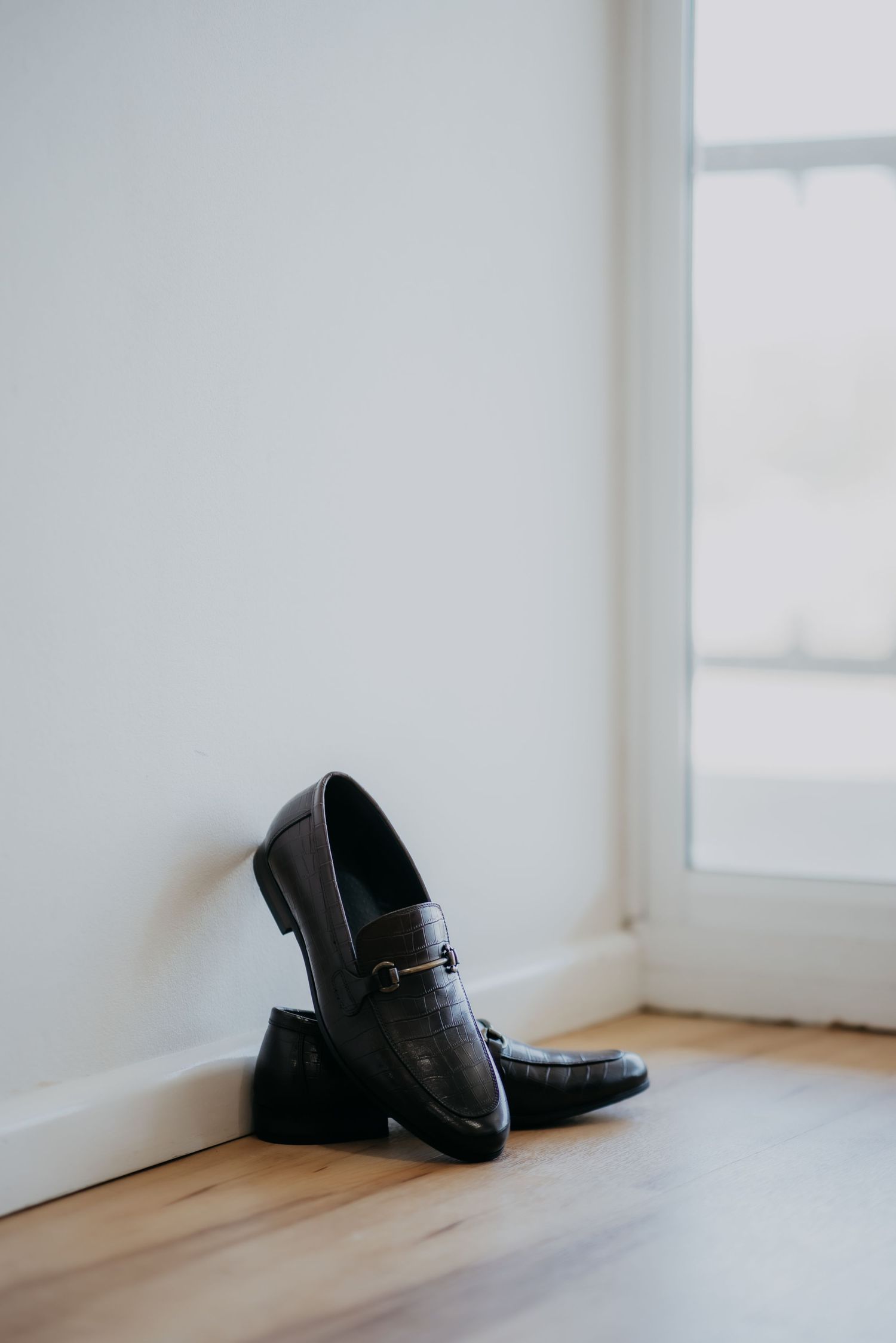Black leather loafer shoes resting against white wall on hardwood floor near window.