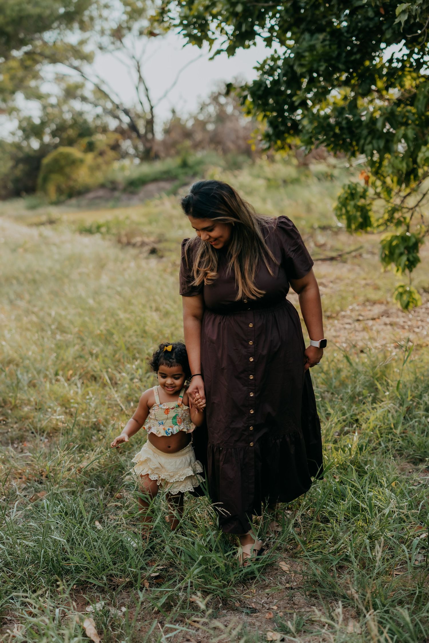 Mother in dark brown dress walking hand in hand with her toddler daughter through a lush green field.