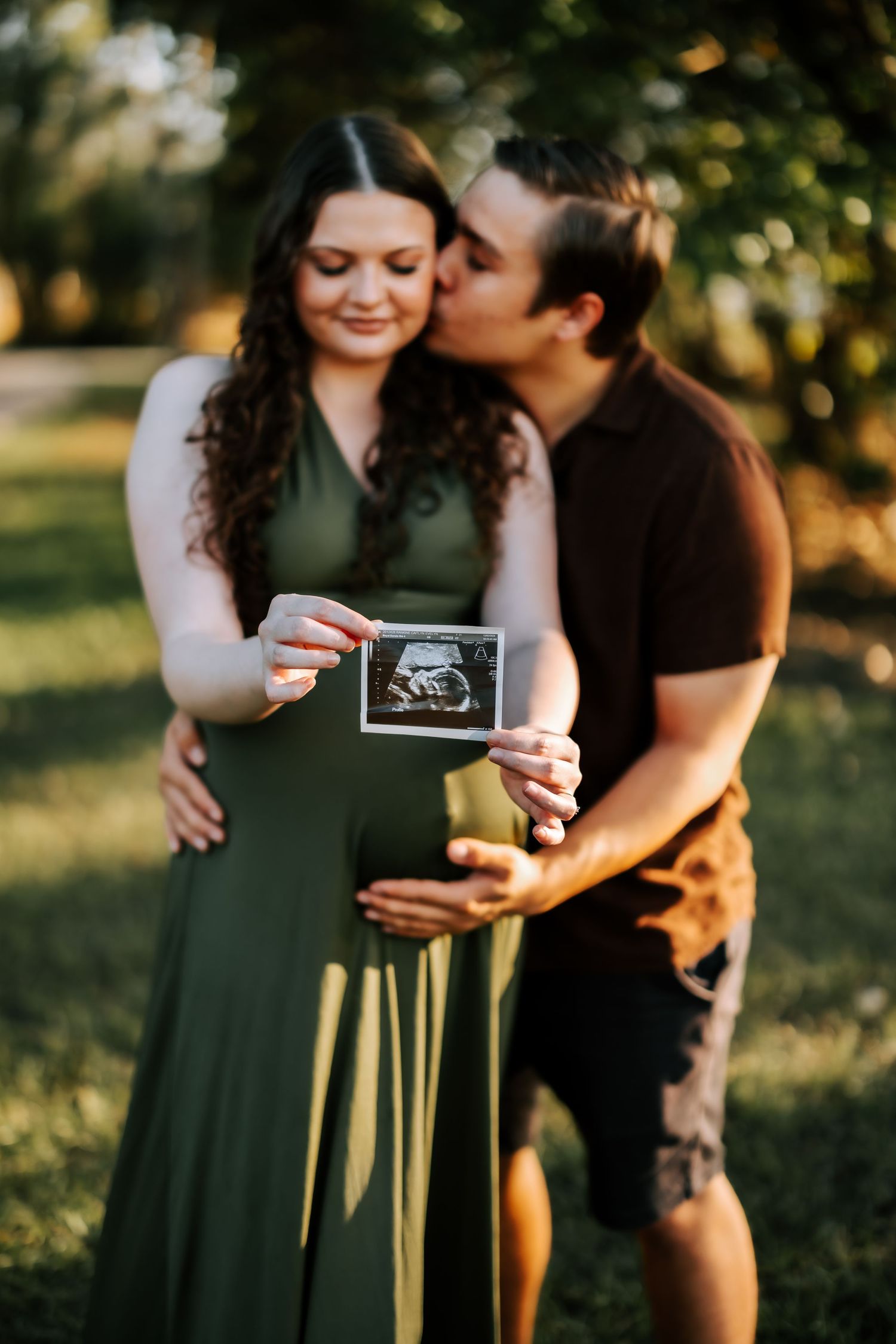Couple holding ultrasound photo in a park, man kissing pregnant woman in green dress.