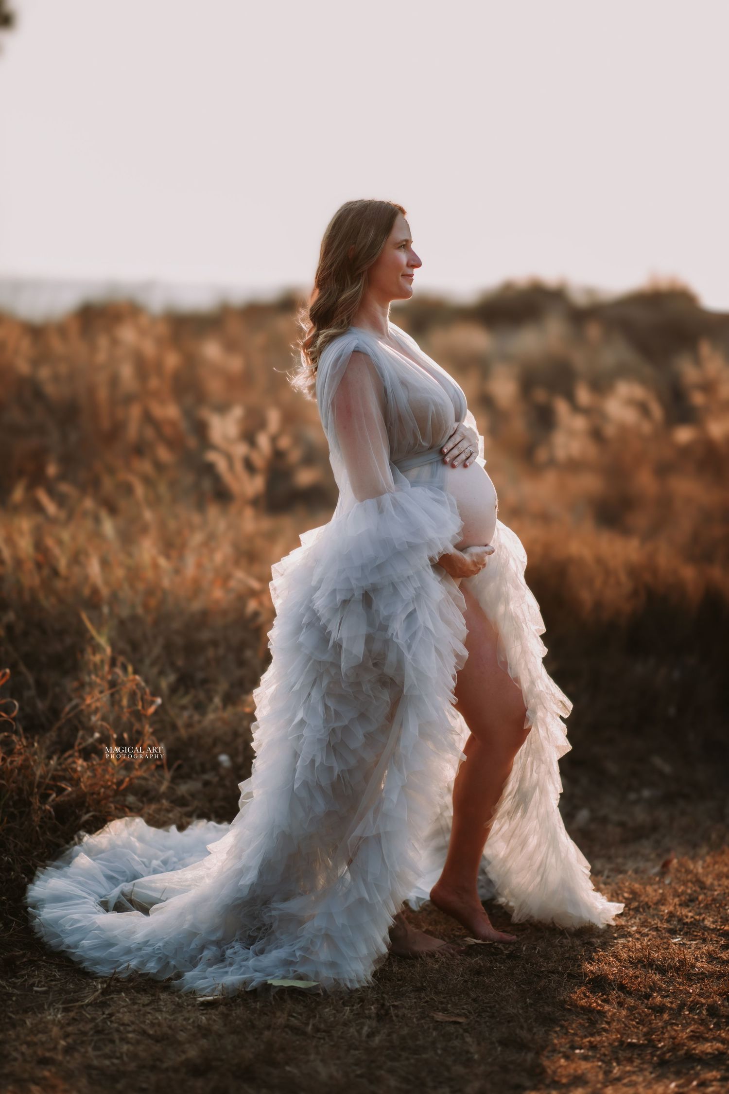 Expectant mother in white ruffled sheer maternity gown posing in golden field at sunset.