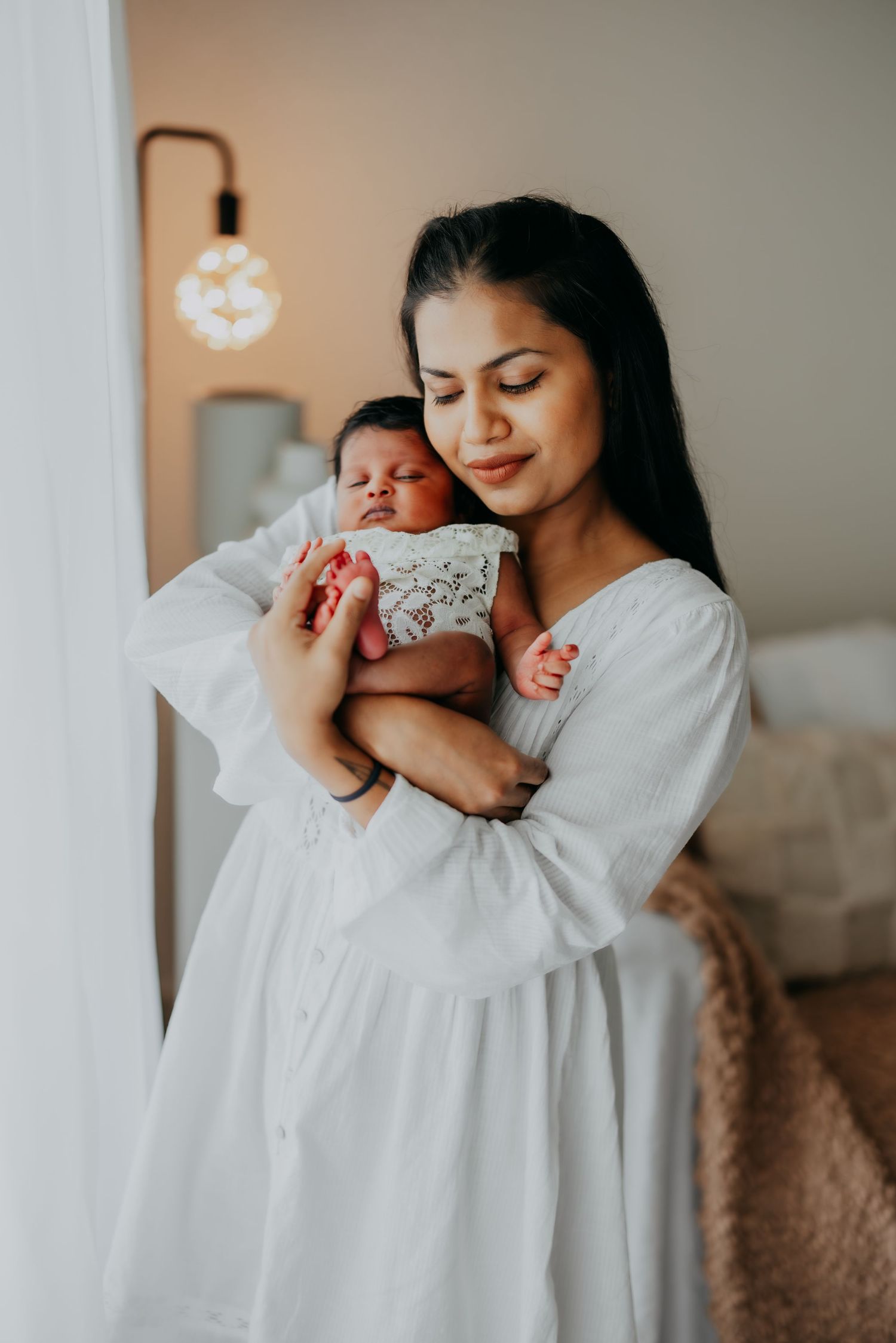 Mother in white dress gently holds sleeping newborn baby indoors.