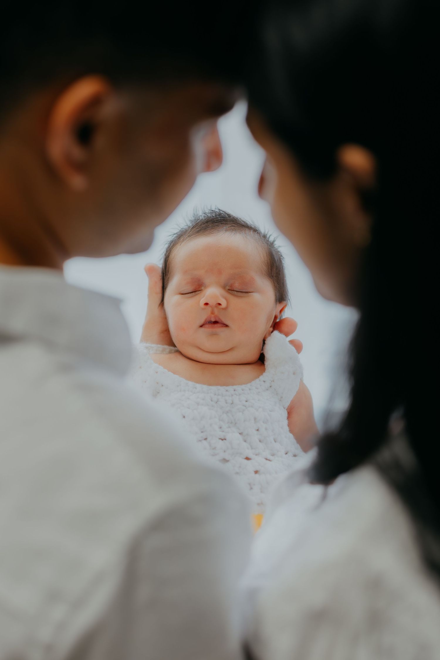 Sleeping newborn baby in white outfit held between two parents seen from behind in soft light.