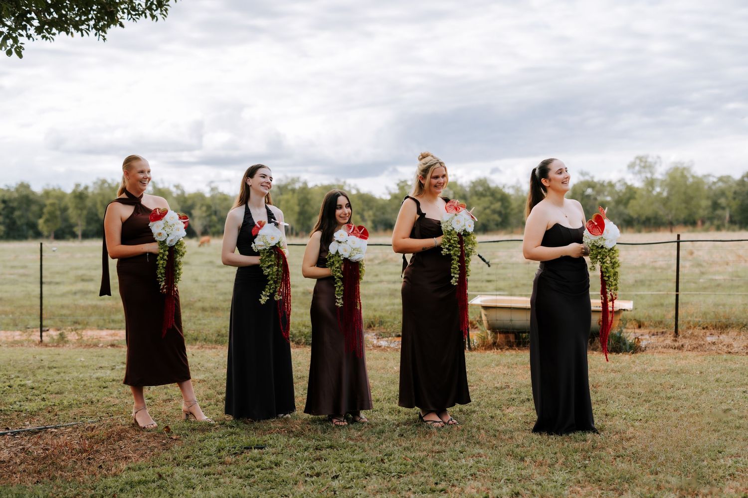 Five bridesmaids in black dresses holding red and white bouquets at an outdoor farm wedding.
