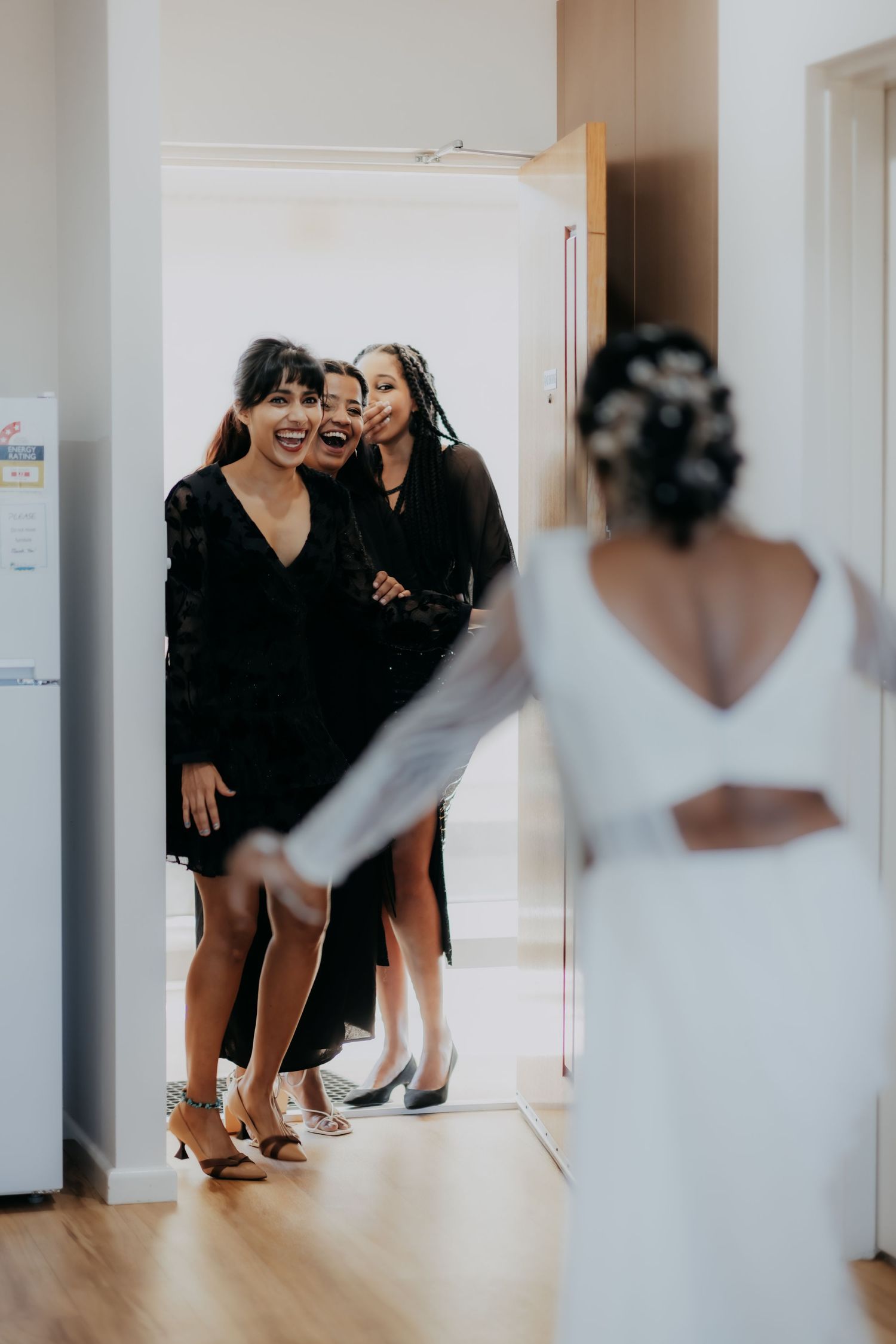 Bridesmaids in black dresses reacting with joy seeing bride in white wedding outfit.