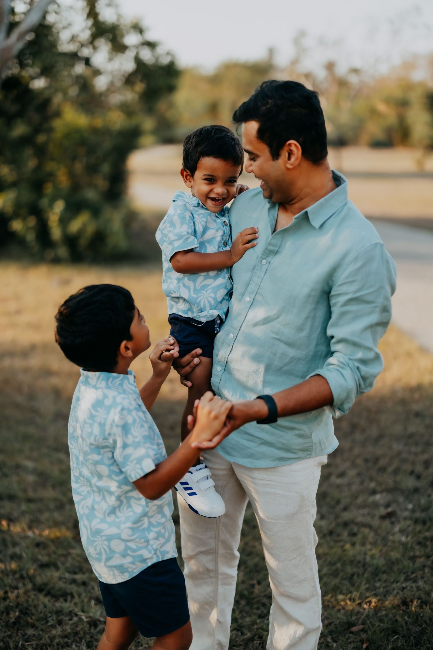 A father in a light blue shirt holds his toddler while his older son smiles up at them outdoors.