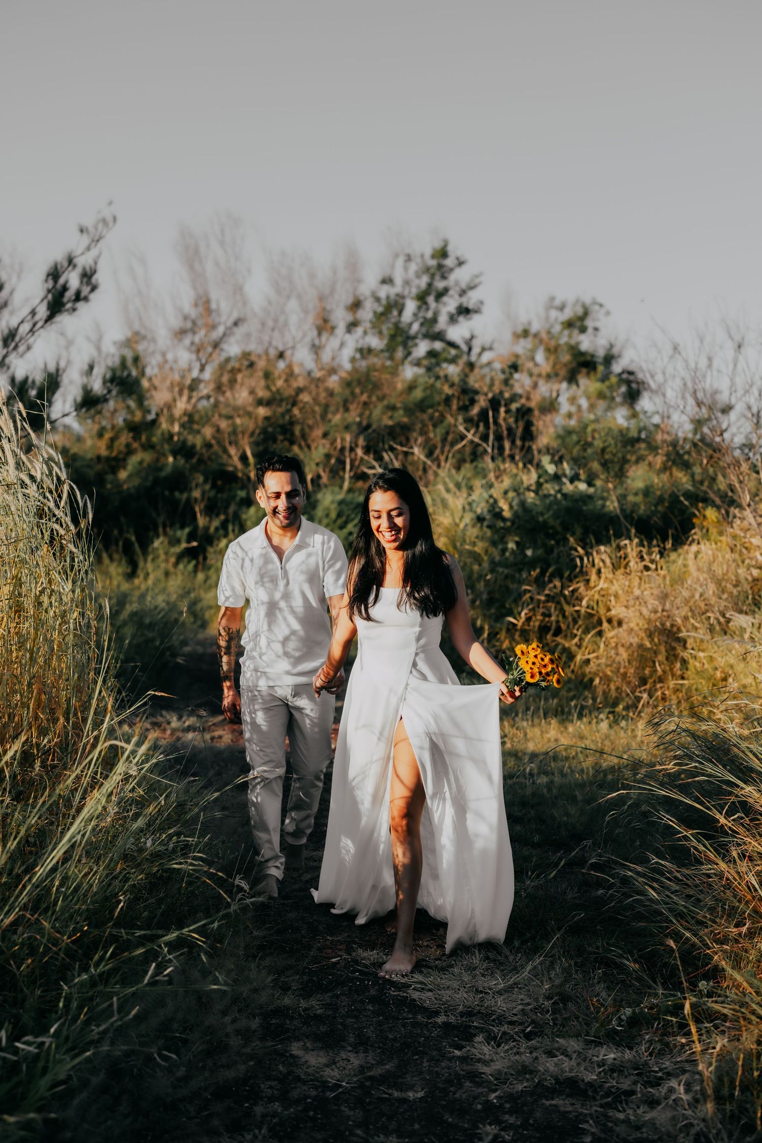 Smiling couple in white outfits walking through a sunlit meadow path, woman holding a bouquet of yellow flowers.