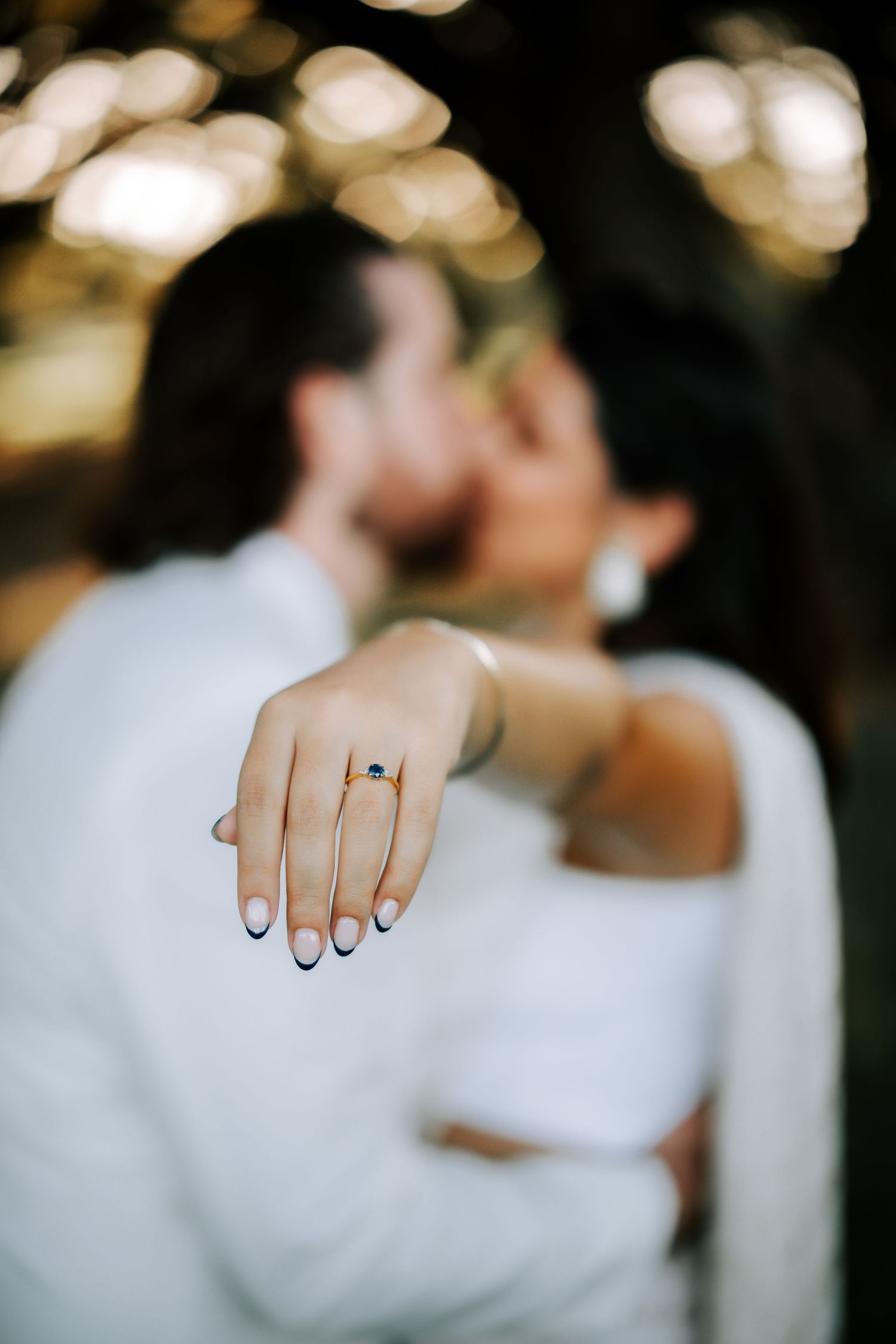 Close-up of engagement ring with dark gemstone on bride's hand while couple kisses in background.