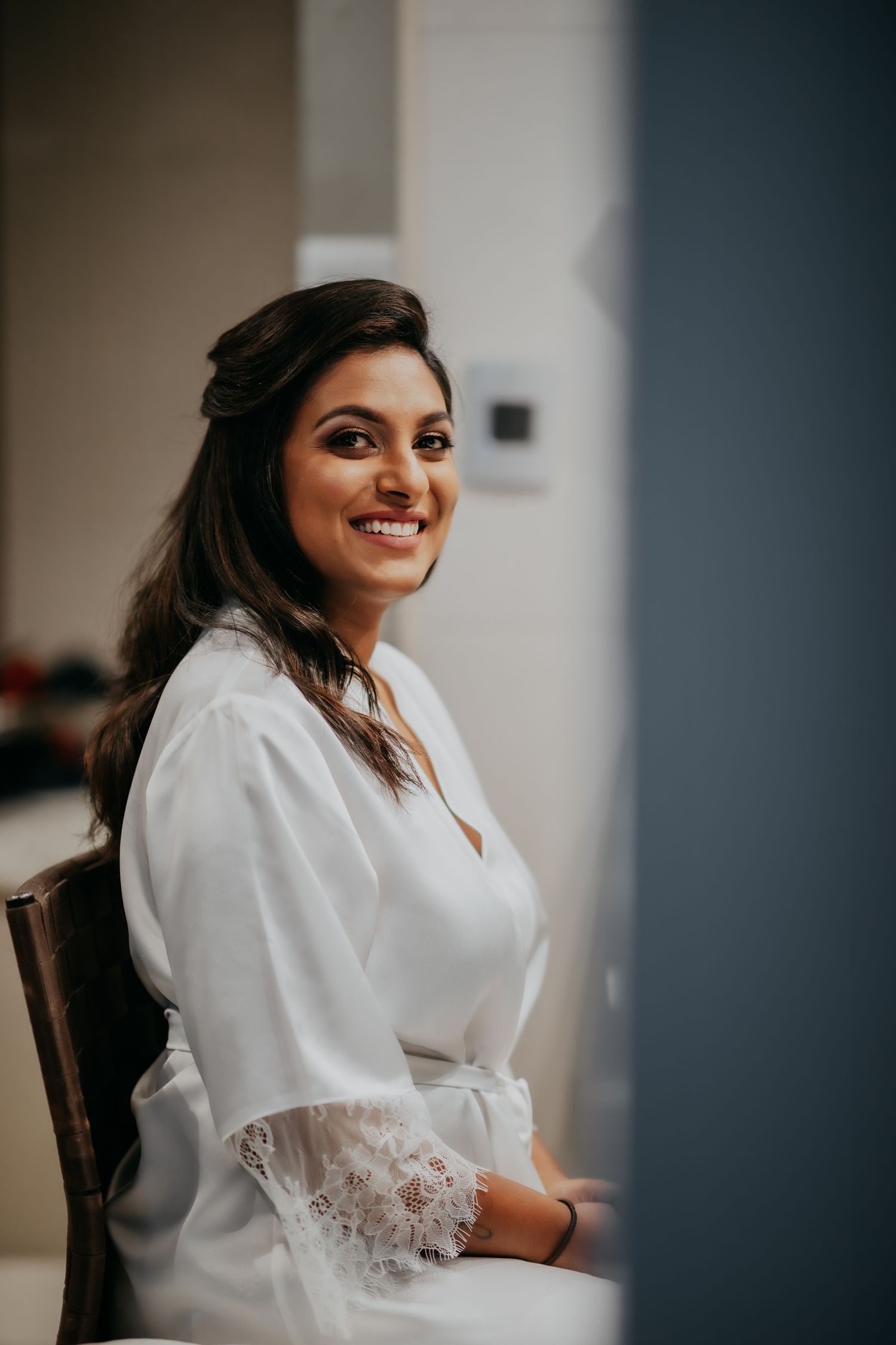 Smiling bride in white lace robe sitting in a chair during wedding preparations.