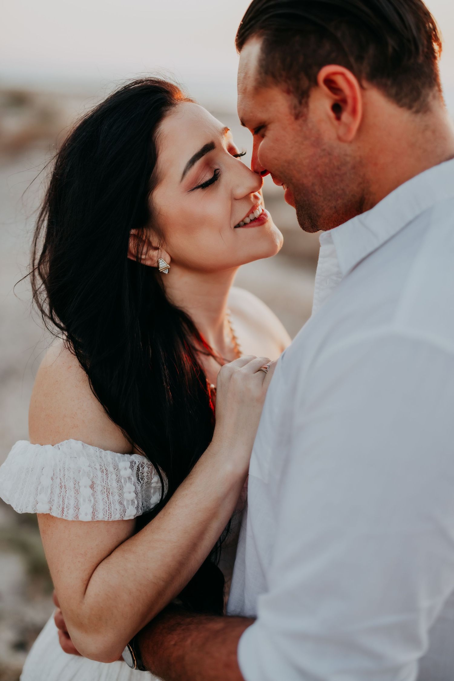 A smiling woman with dark hair leans close to a man as they almost kiss in warm sunset light.
