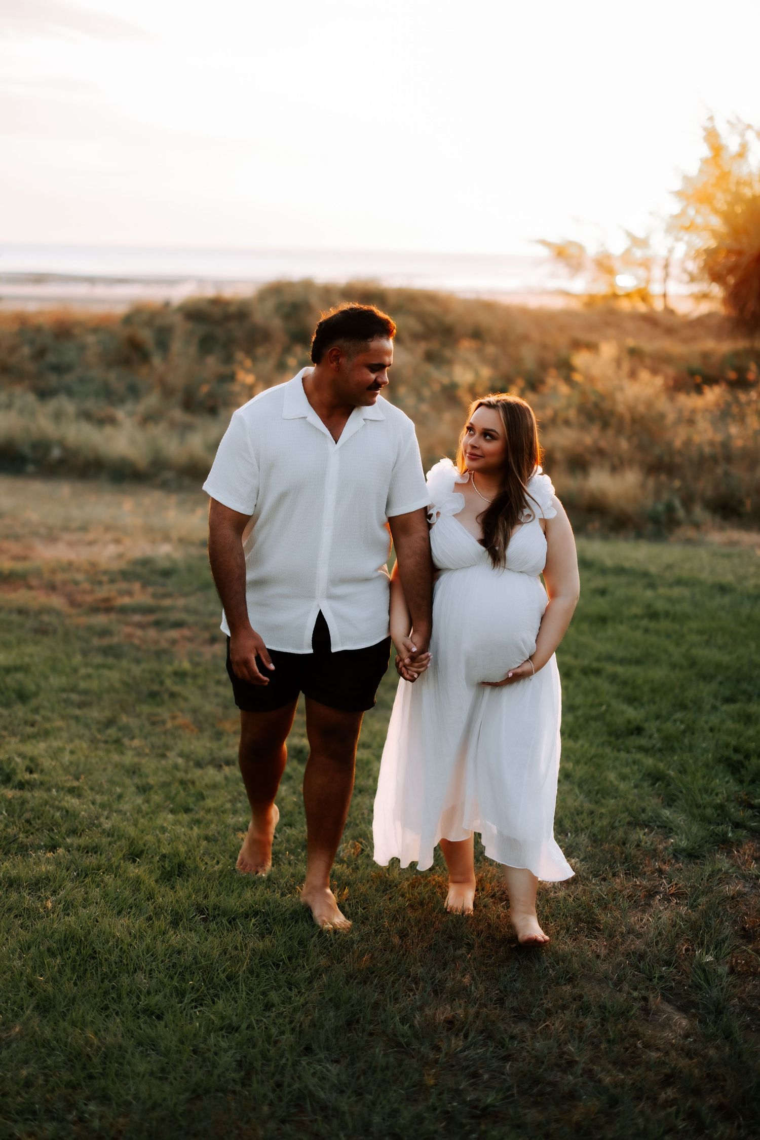 Pregnant couple walking hand-in-hand in grassy field at golden hour sunset.