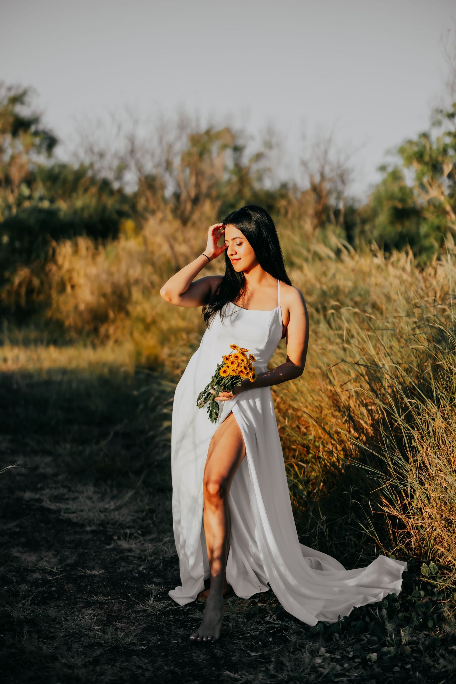 Elegant woman in white slit dress holding sunflowers, posing barefoot in golden field at sunset.