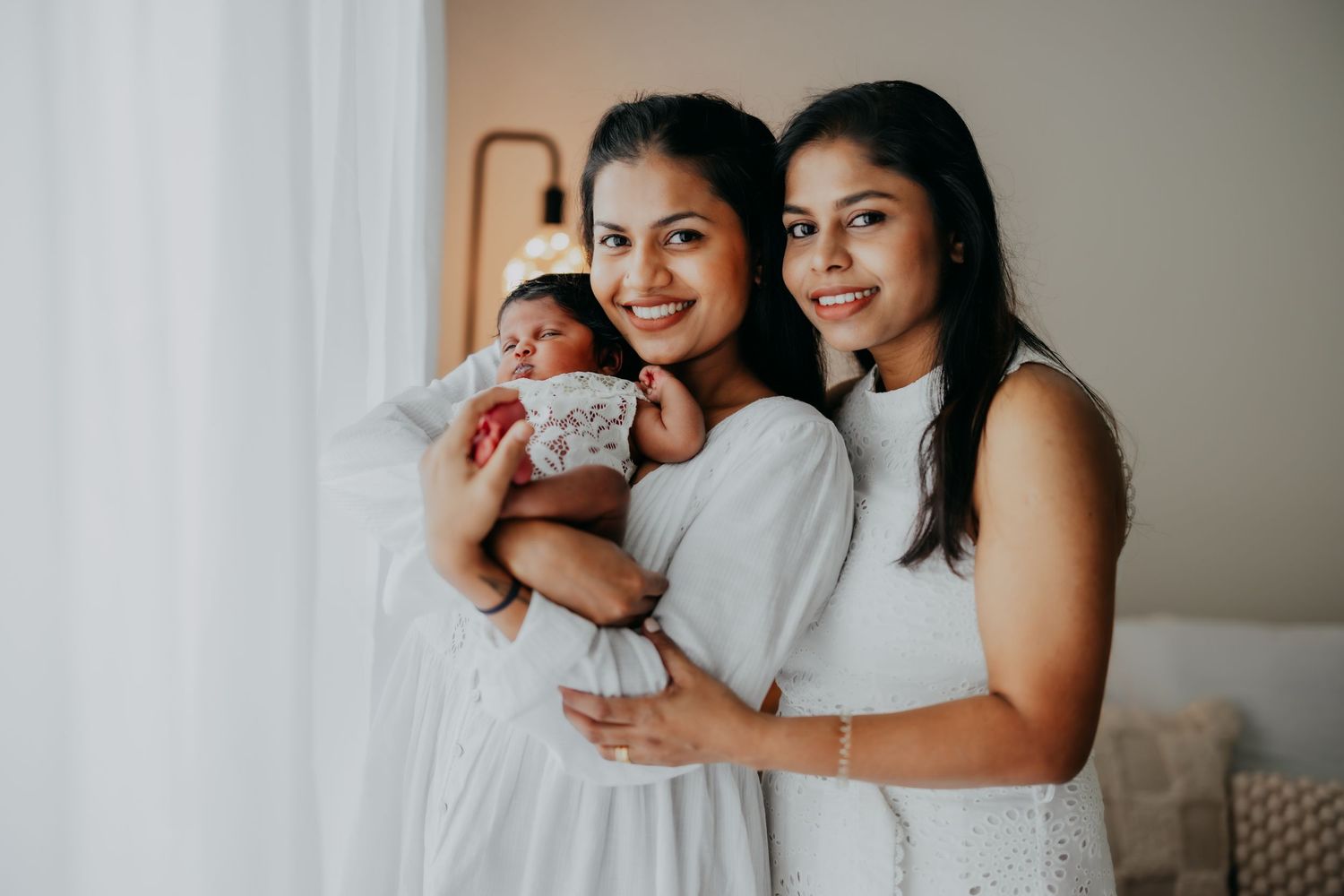 Two women in white dresses pose with a newborn baby by window.