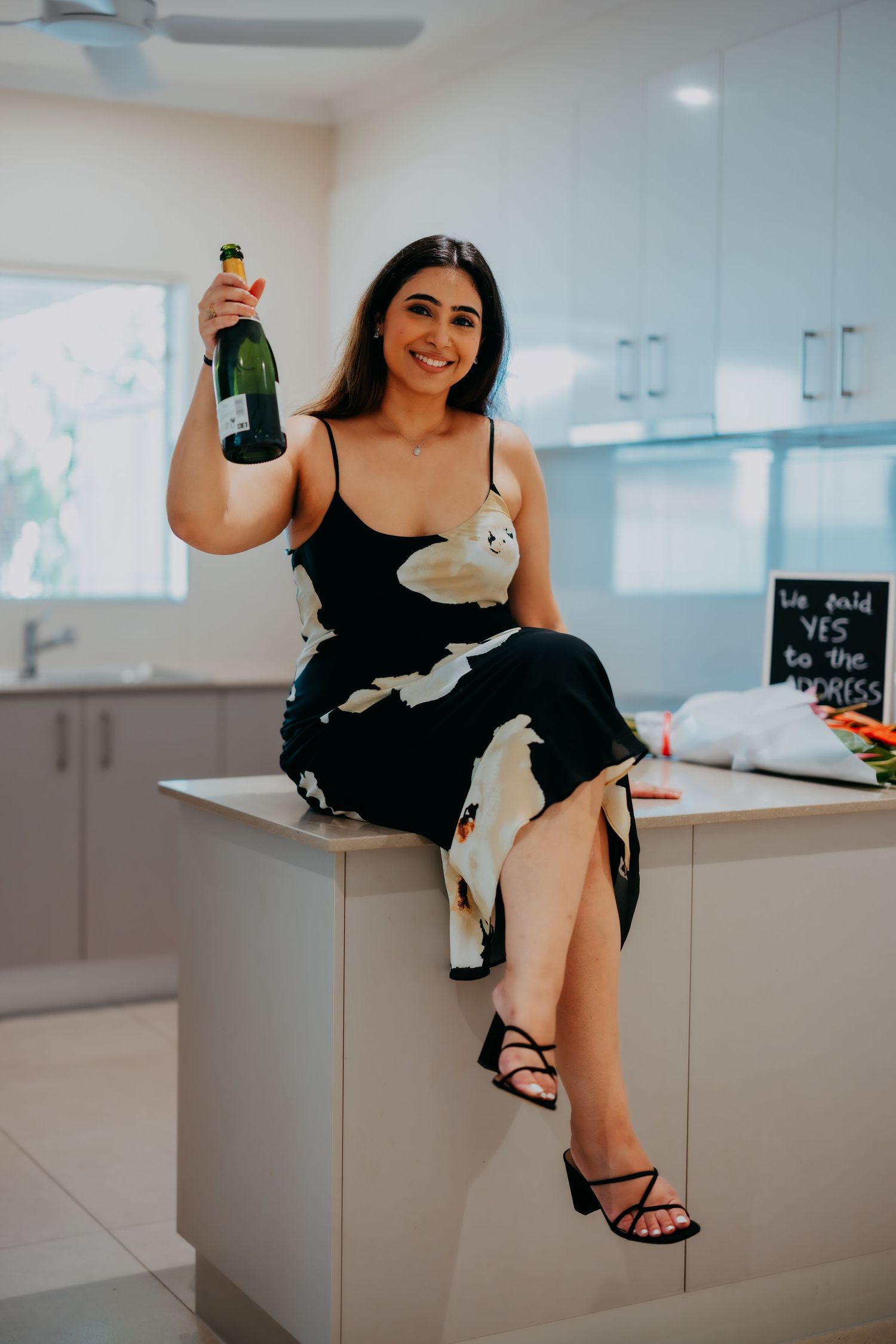 Woman in black floral dress sits on kitchen counter holding champagne bottle, smiling.