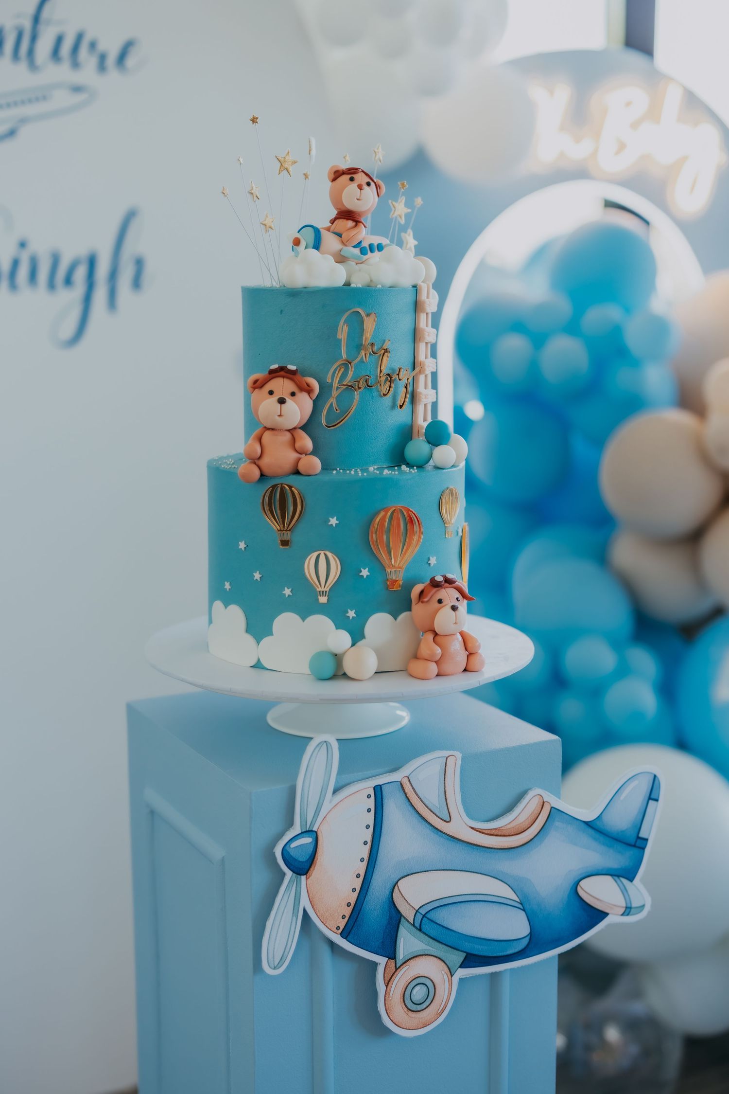Blue two-tier baby shower cake with teddy bear toppers, hot air balloons, and airplane decorations surrounded by blue balloons.