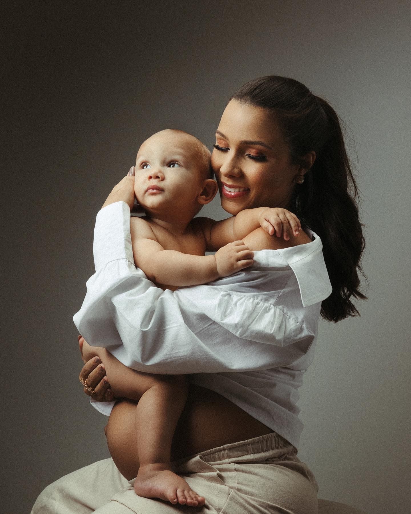 Tender mother and baby portrait session with white outfits against grey backdrop.