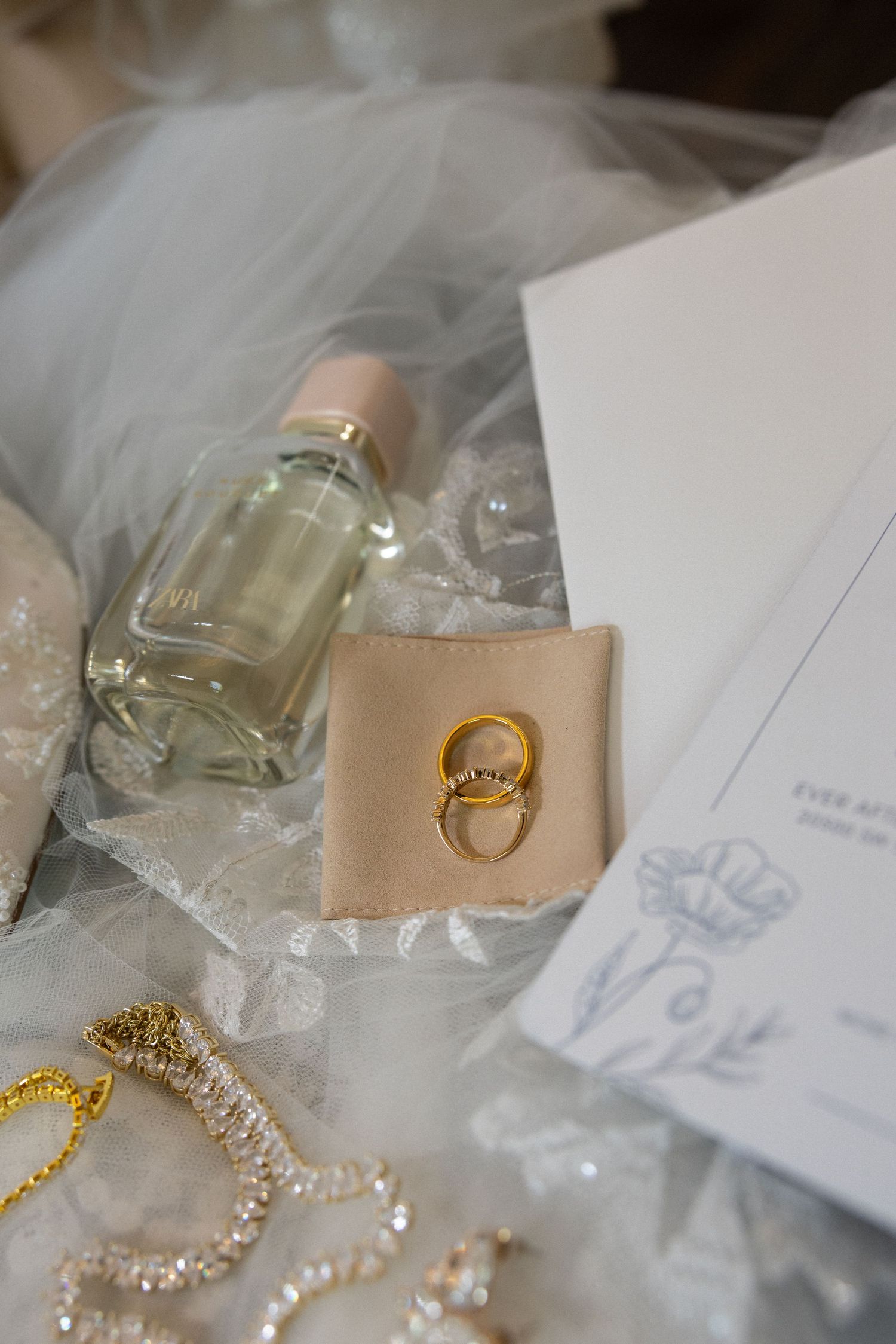 Wedding accessories including perfume bottle, gold ring on beige leather, and delicate pearl jewelry with tulle fabric.