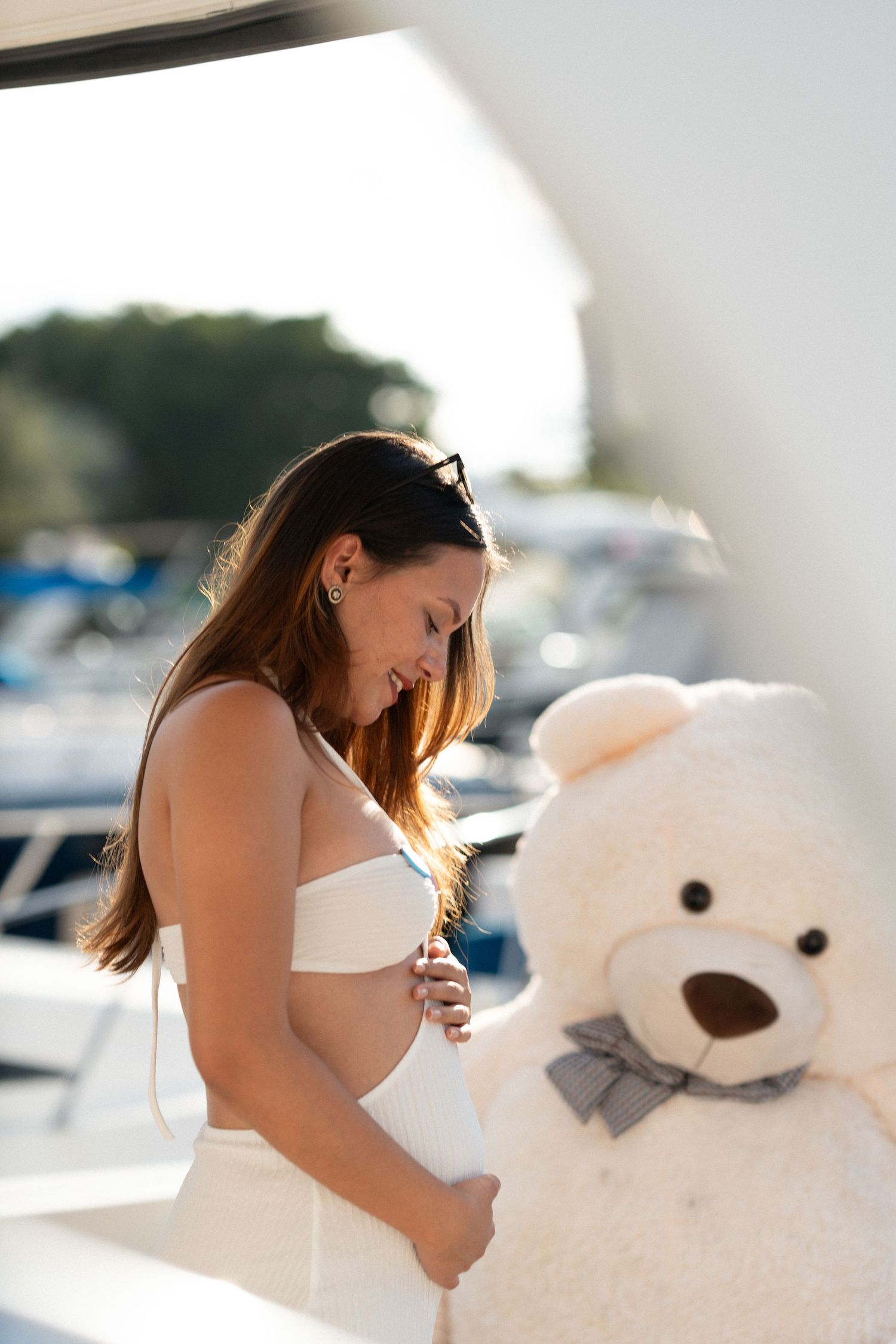A series of photos showing a pregnant woman in white embracing a large white teddy bear outdoors near a marina.