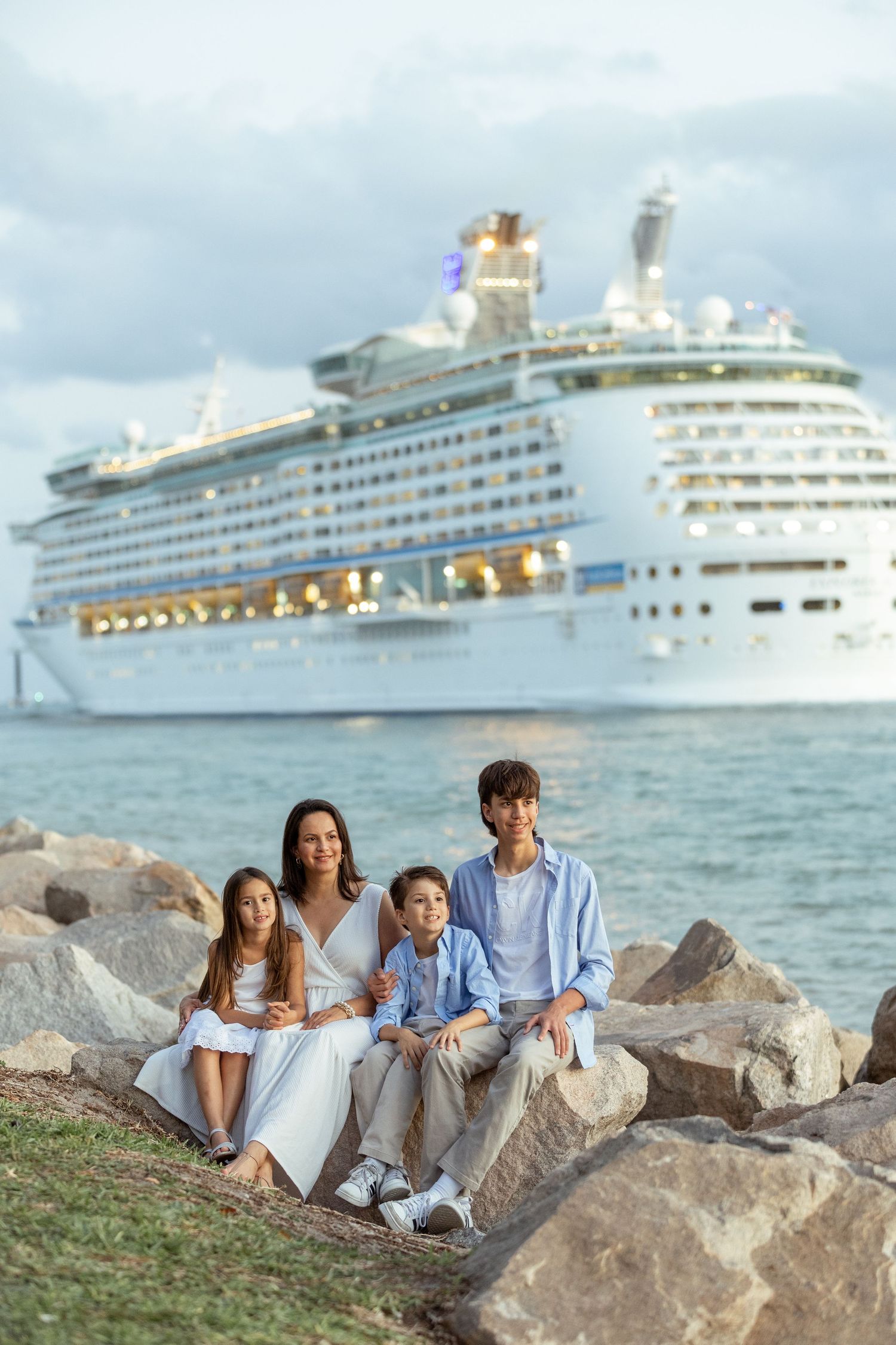 Family relaxing on rocky beach with luxury cruise ship in background during sunset.