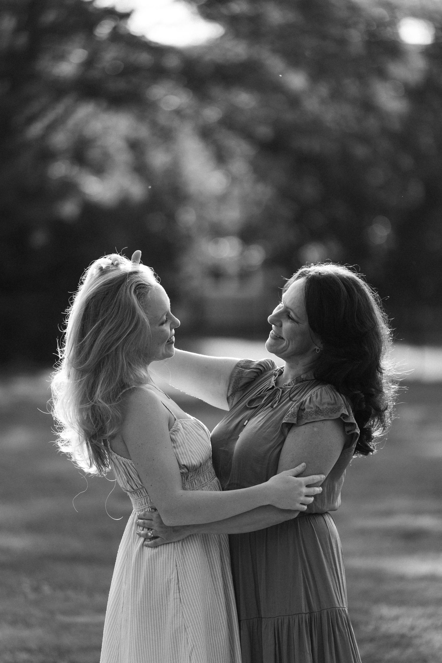Two women who are mother and daughter in flowing dresses share a joyful embrace in a sunlit outdoor setting, captured in black and white.