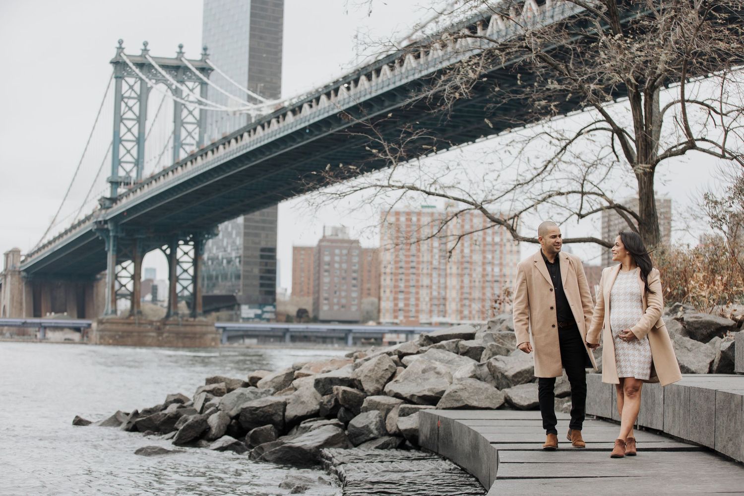 Couple walks along rocky shoreline under Manhattan Bridge in Brooklyn with NYC skyline in background.