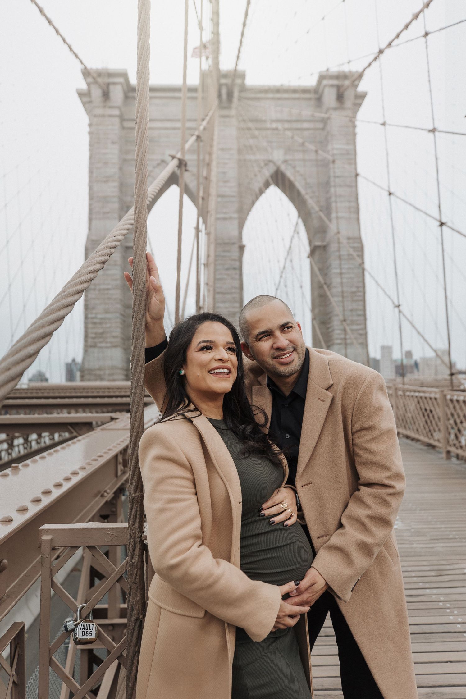 Stylish couple in camel coat and black dress posing on Brooklyn Bridge.