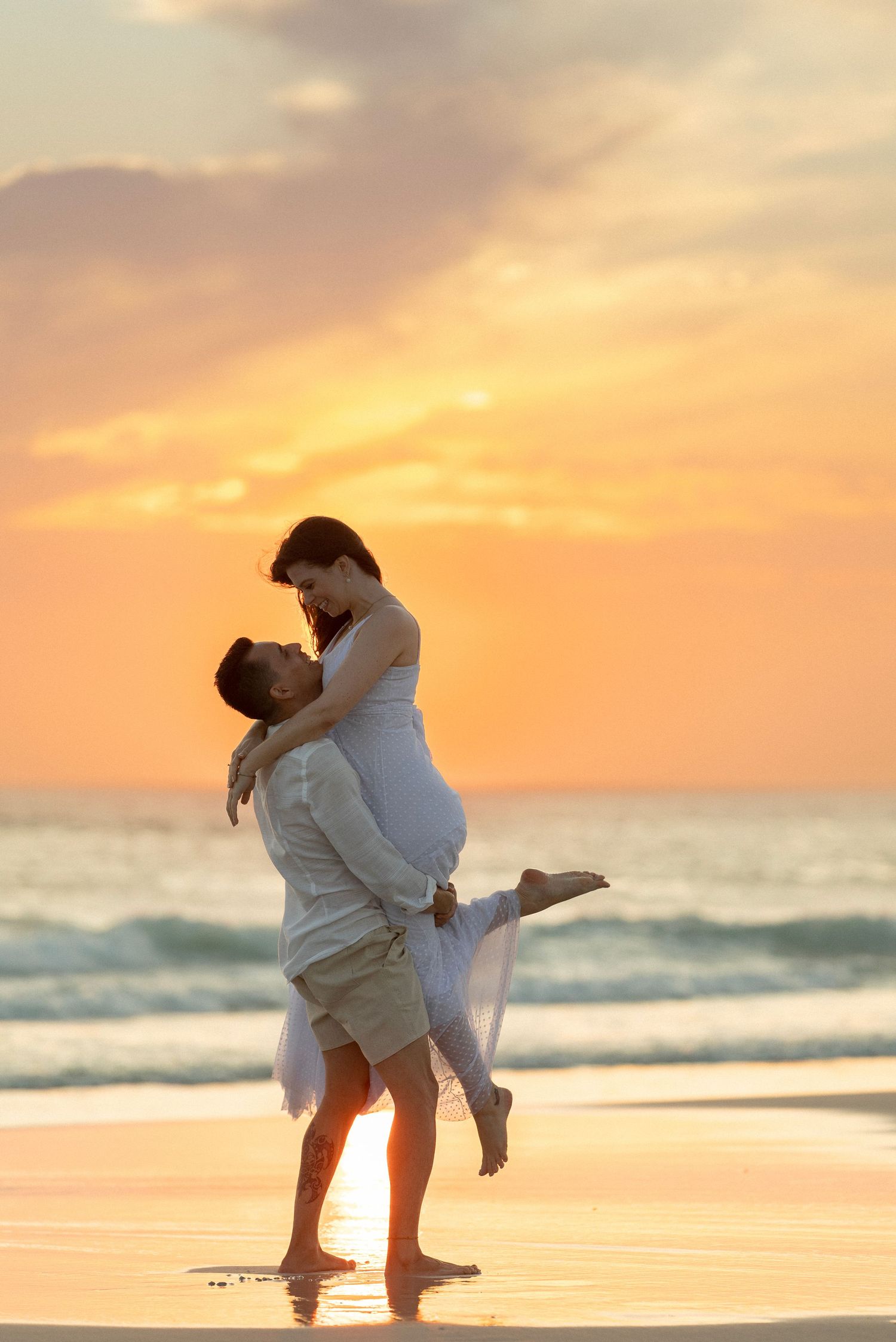 Romantic beach photoshoot at sunset with couple in white attire.