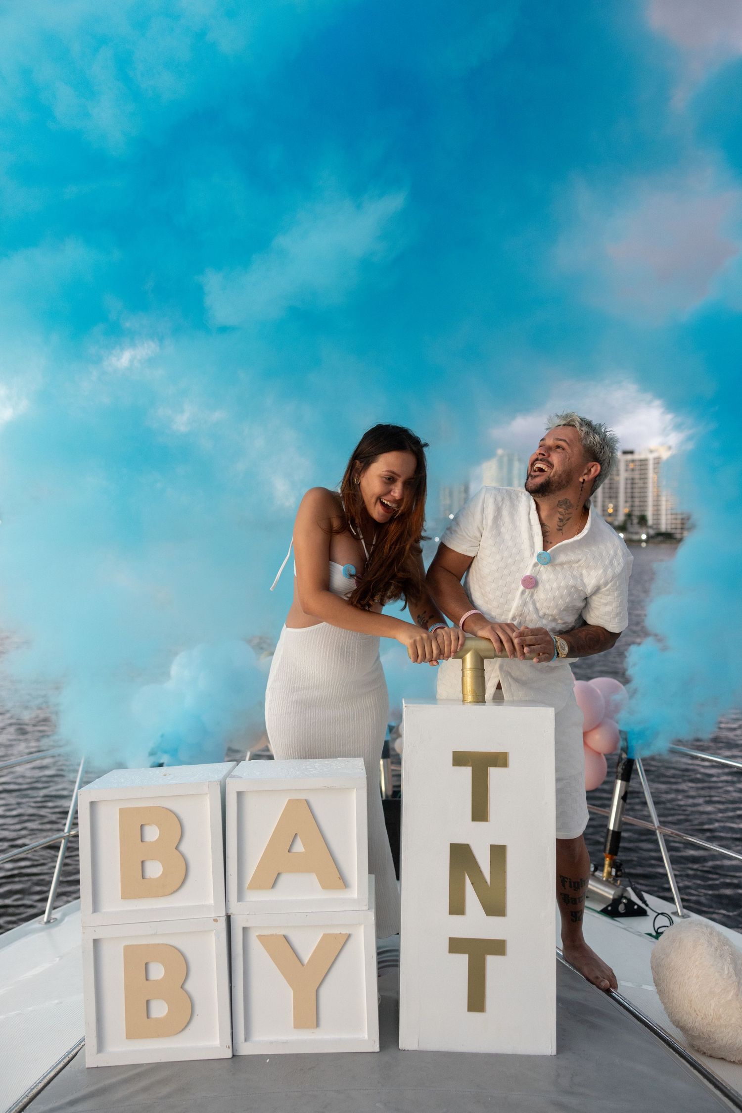A creative pregnancy announcement photo series spells out 'BABY IT' using white letter blocks on a dock with blue sky.