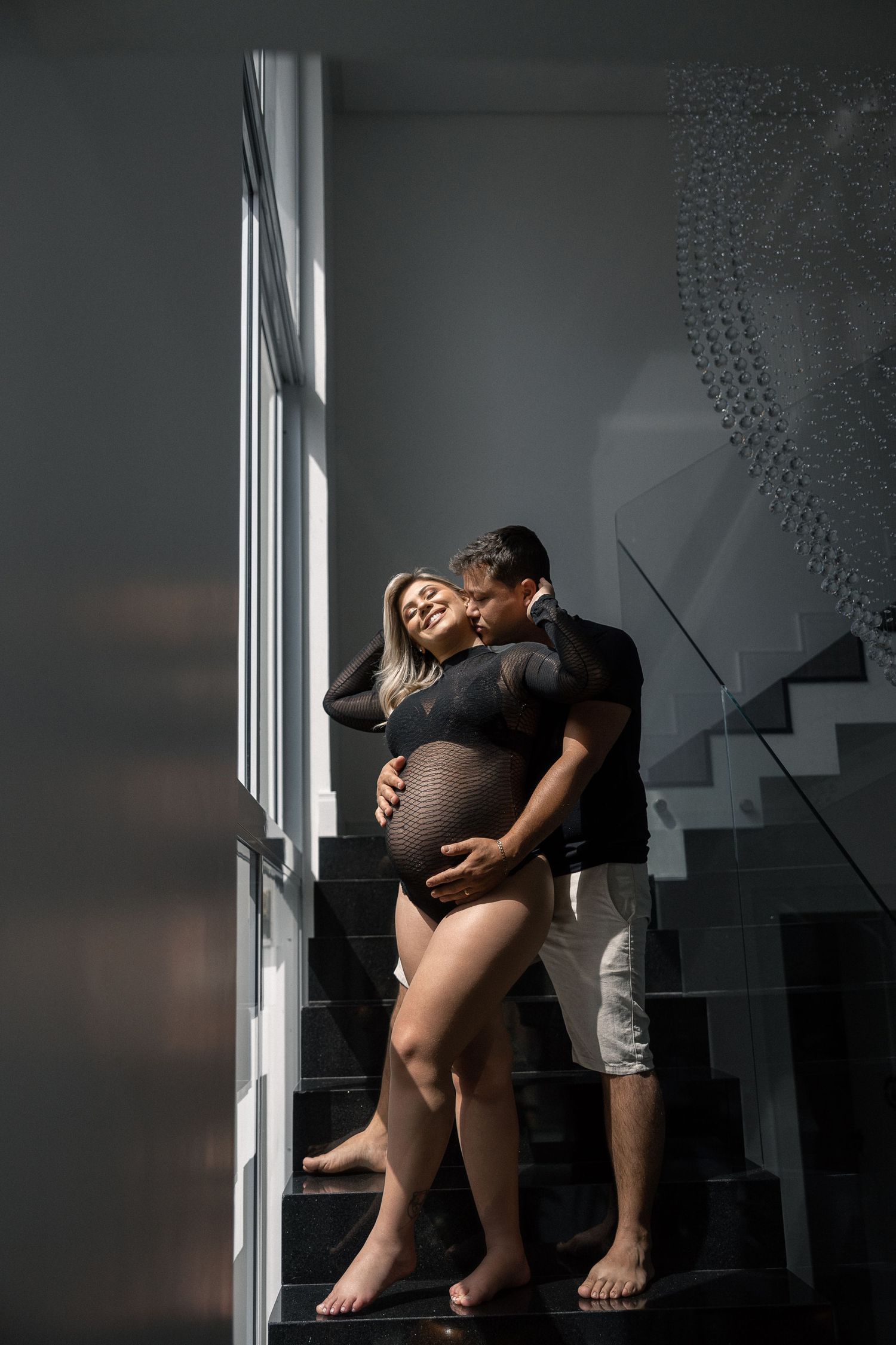 A couple shares an intimate moment on dark stairs near a window in a maternity photoshoot setting.