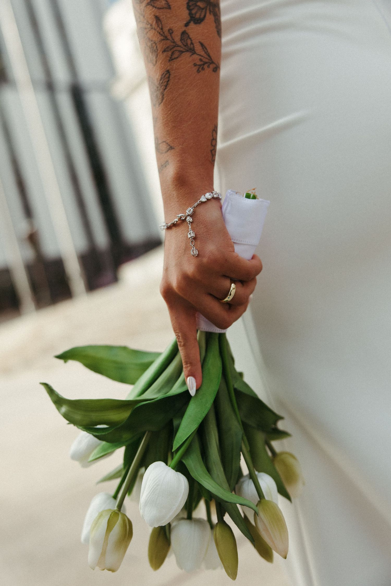 Close-up view of hands holding elegant white tulips against a white dress with delicate jewelry and tattoo details.