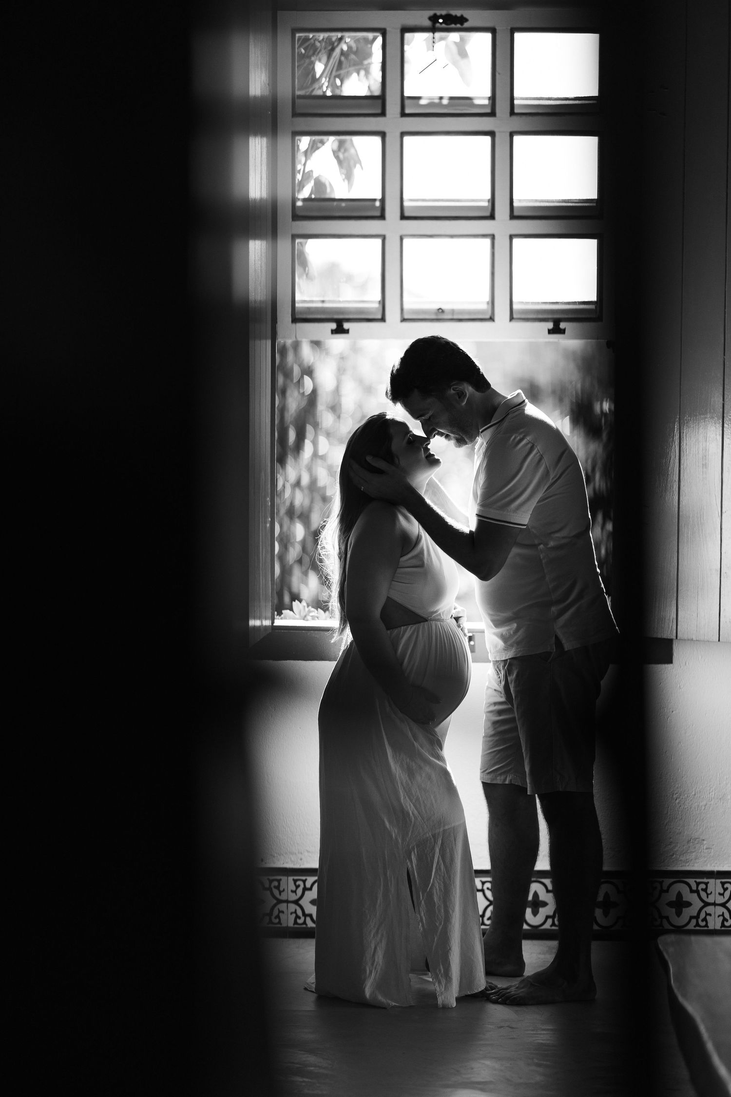 Silhouetted figures share an intimate moment against a bright window backdrop in black and white photography.
