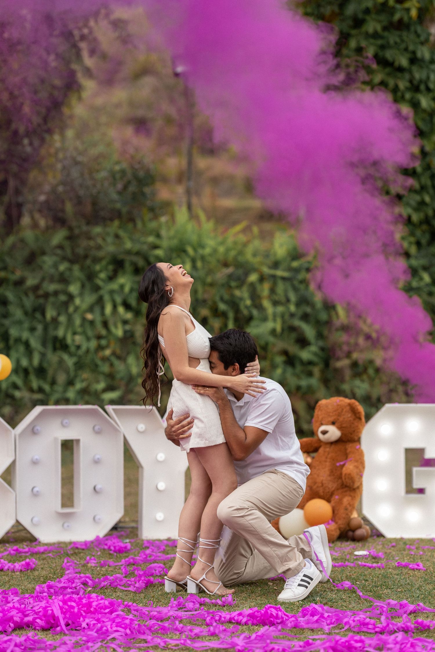 A gender reveal celebration with pink smoke and illuminated letters in an outdoor setting.