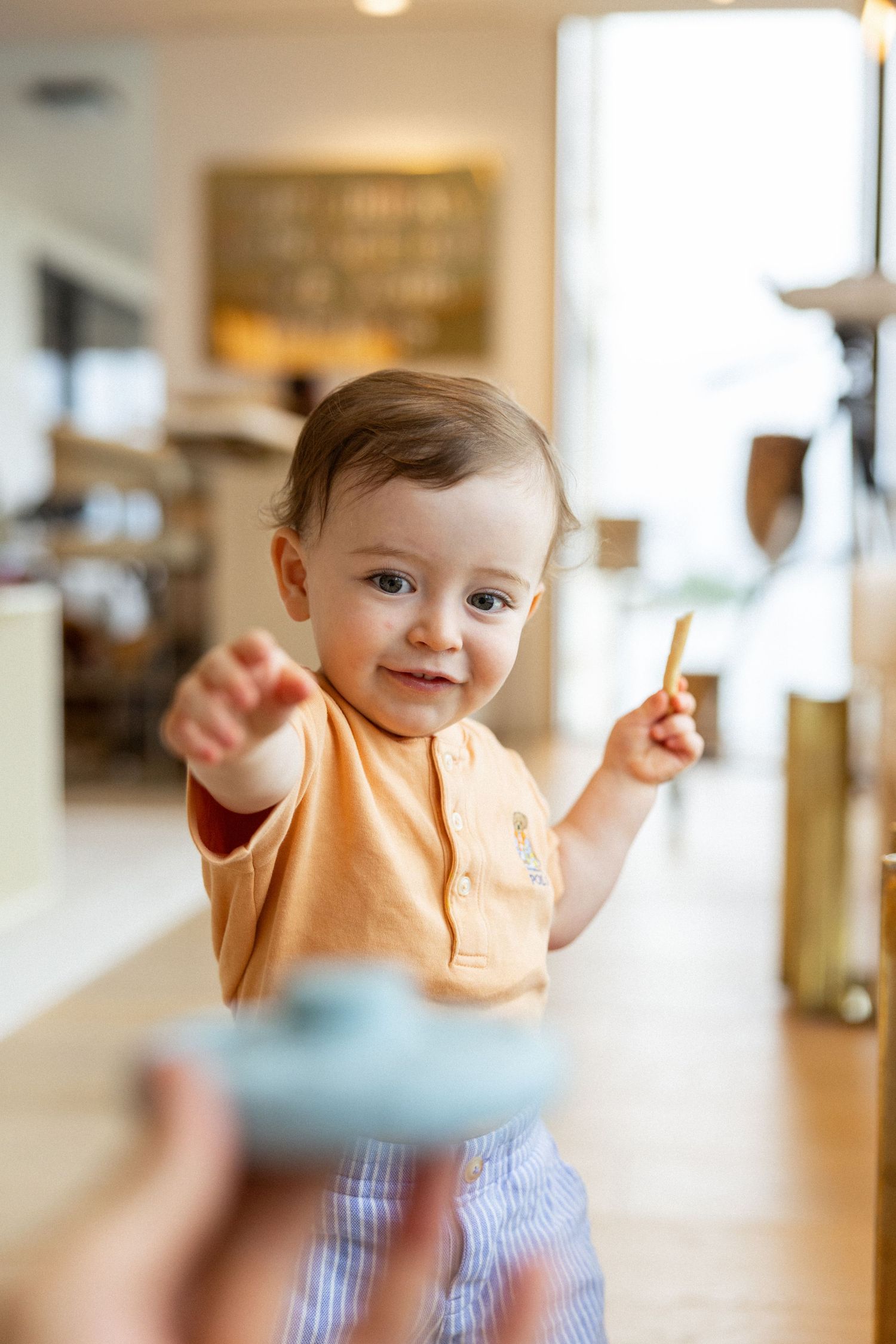 Baby in peach colored shirt and blue gingham pants plays and gestures in a bright indoor space.