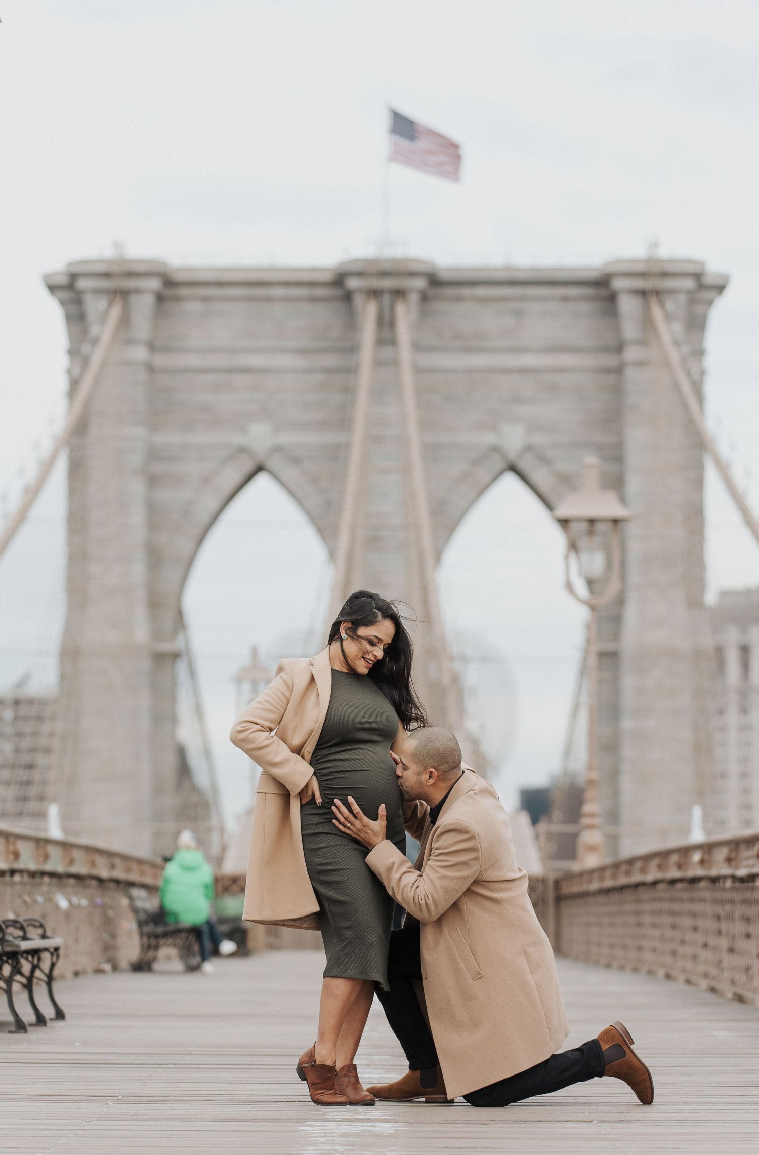 A tender pregnancy moment captured on the Brooklyn Bridge with an American flag waving in the background.