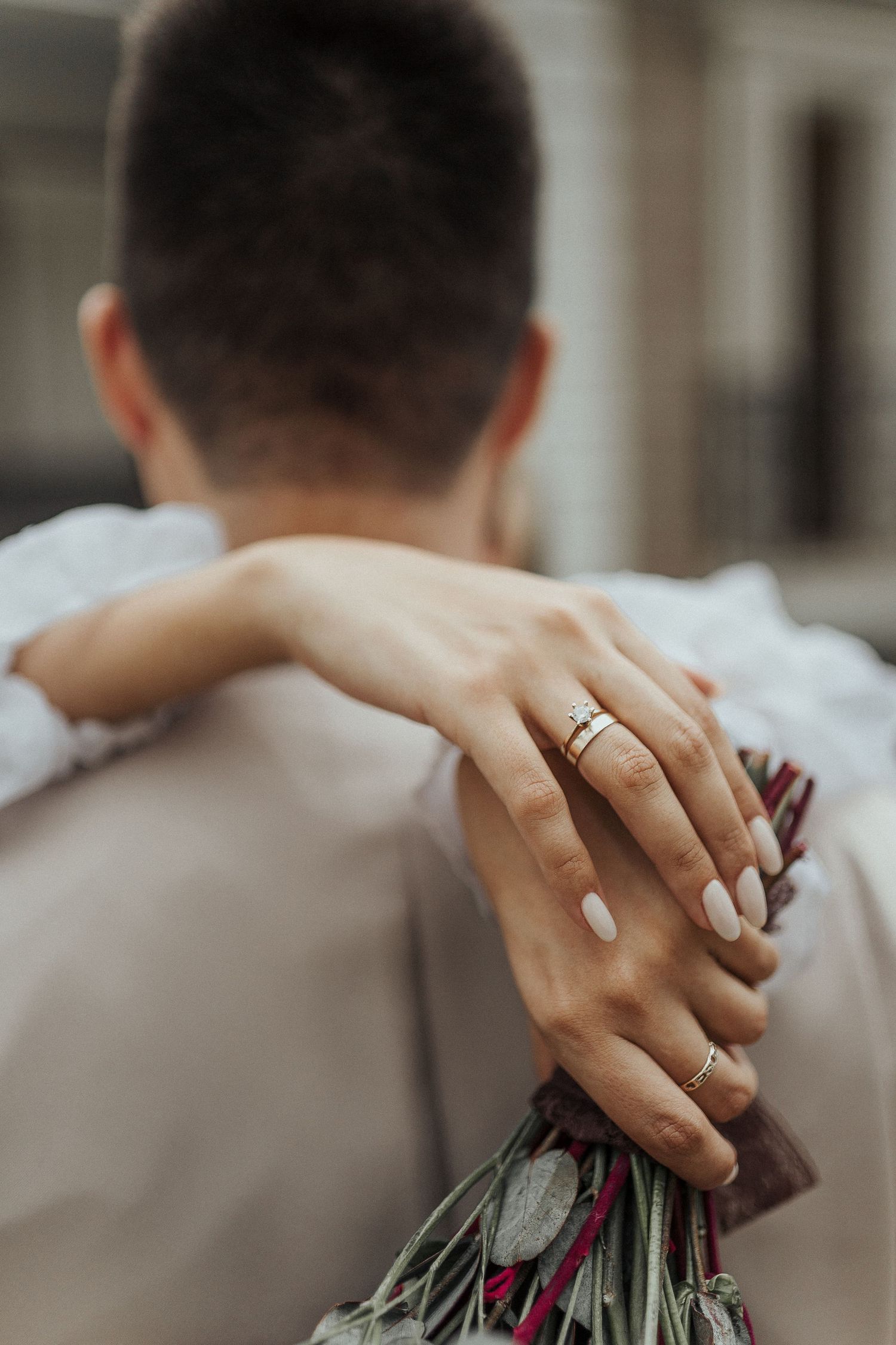 Close-up of hands with wedding rings holding pink flowers against a neutral background.