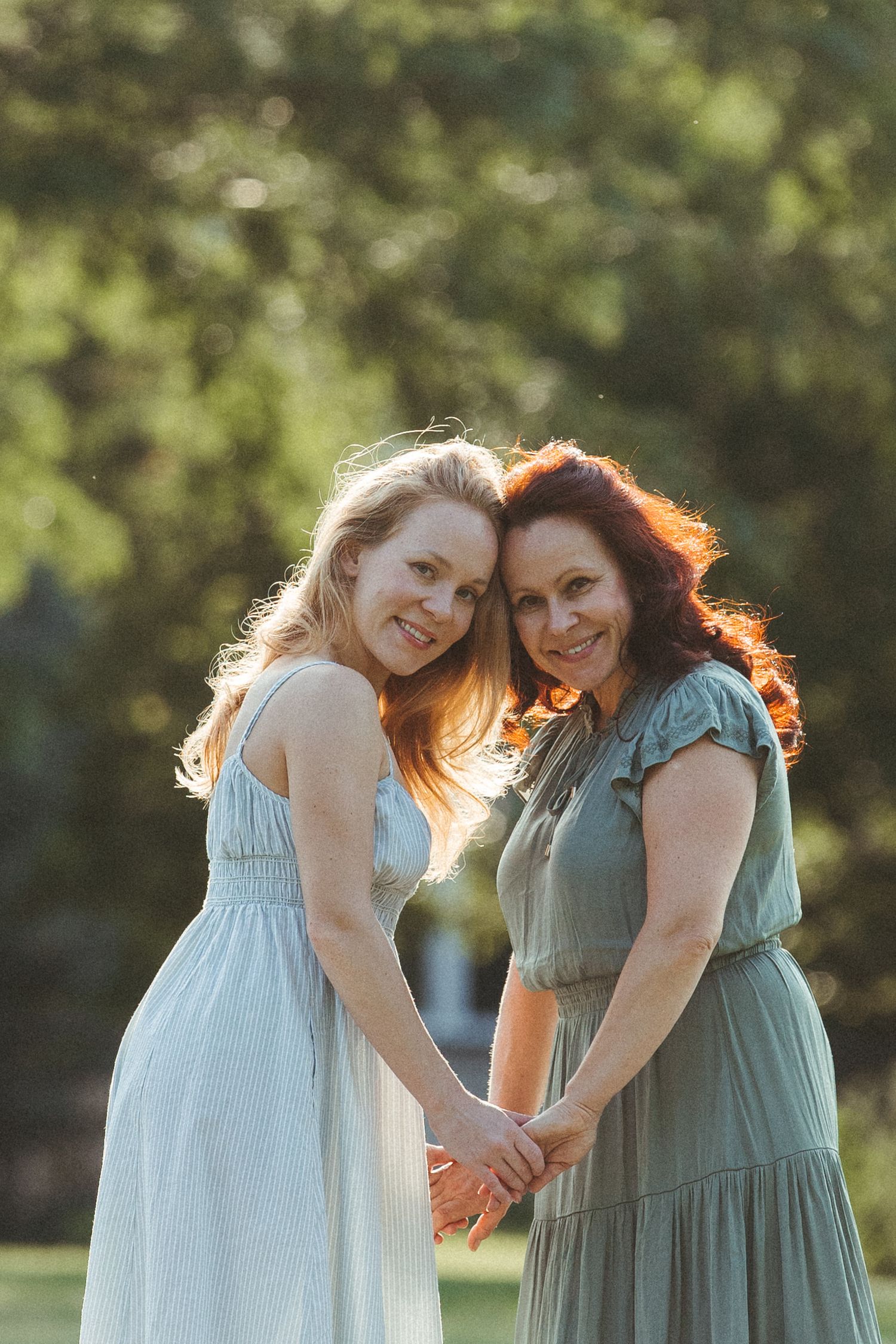 Two women in light summer dresses pose together outdoors with golden sunlight illuminating their hair.