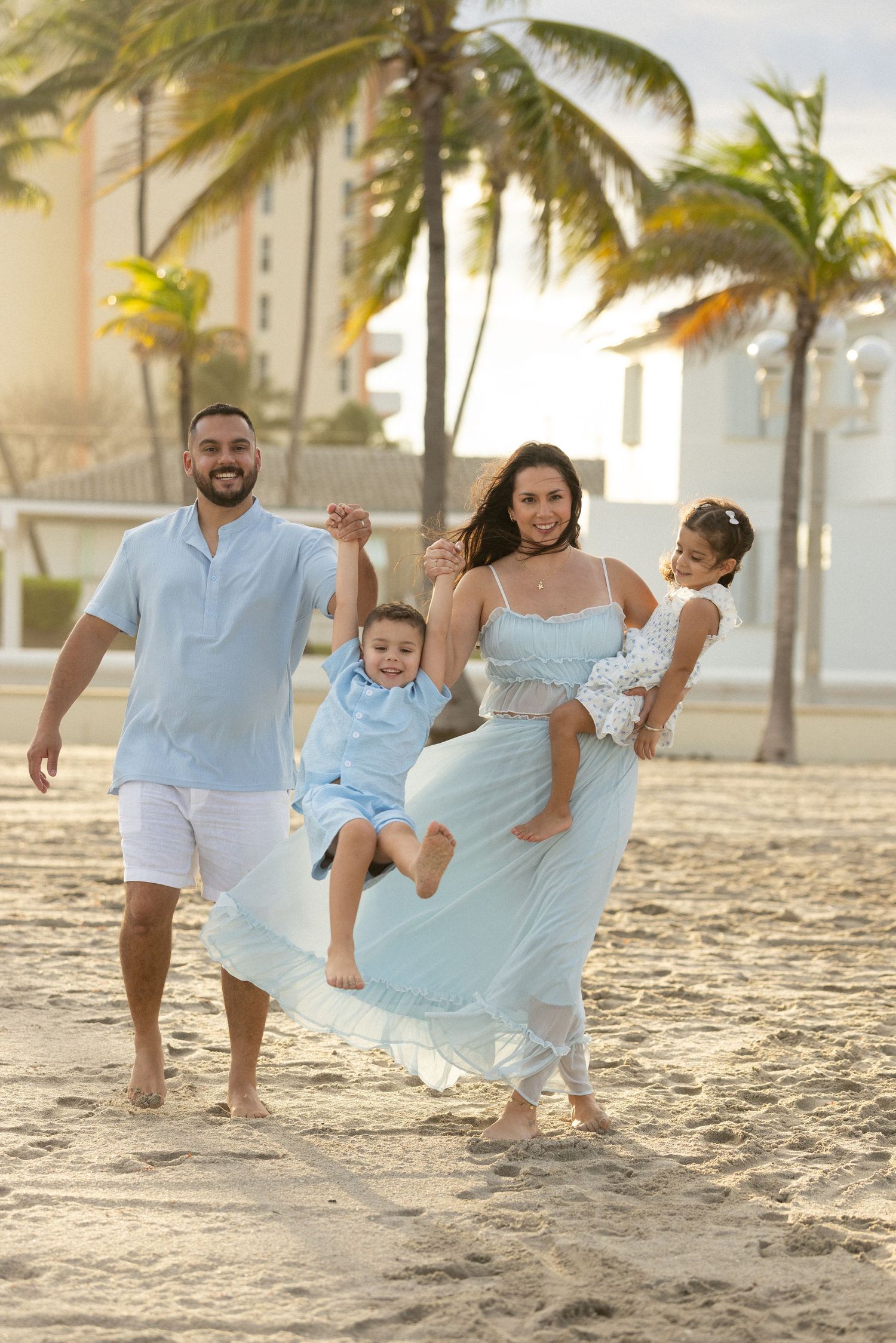 Happy family plays together on a sandy beach with palm trees during golden hour sunset.
