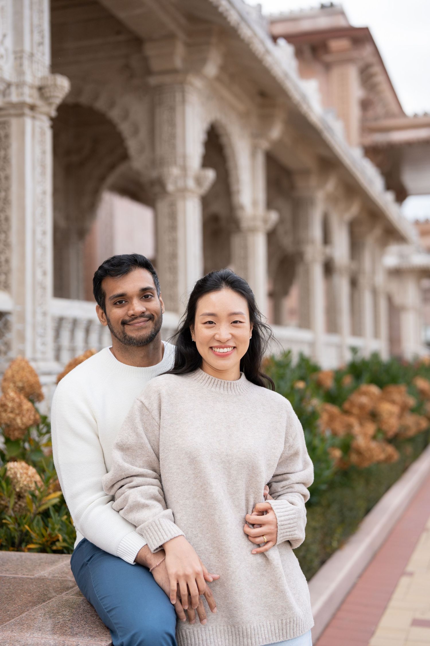 A couple in matching cream sweaters sits together on stone steps with ornate architectural columns in the background.