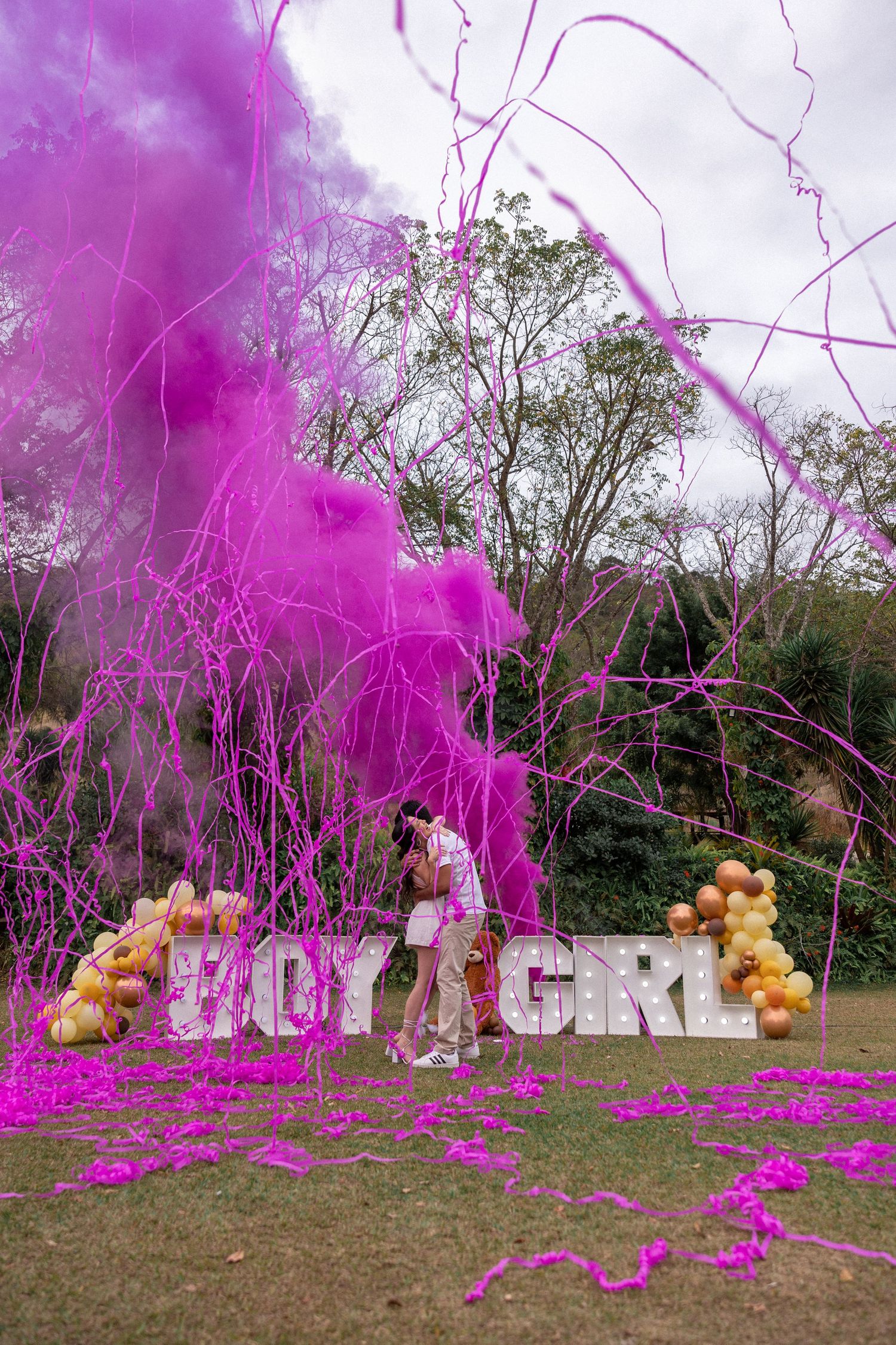 Gender reveal celebration with vibrant pink smoke bombs spelling out text in an outdoor grassy field.