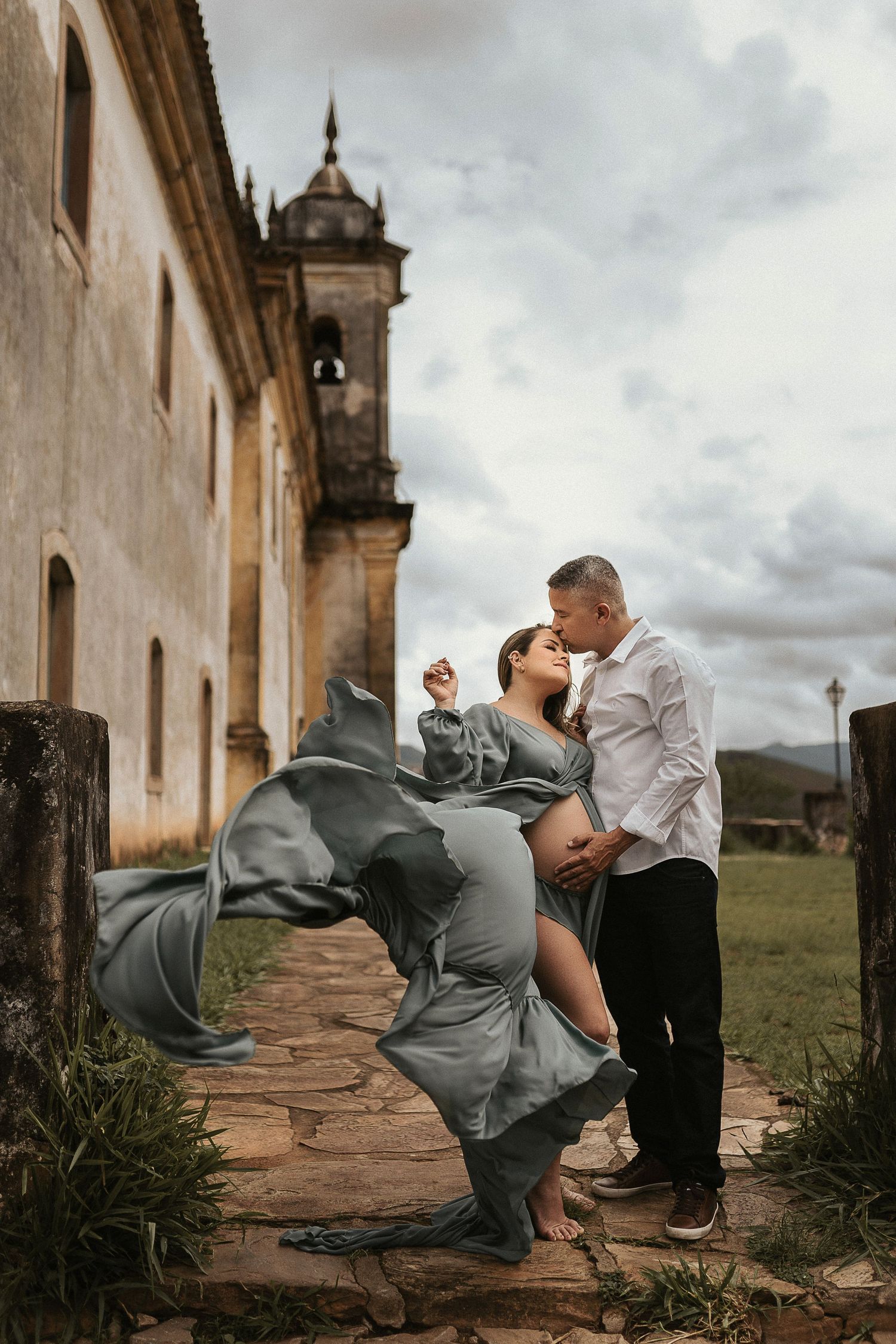 A romantic couple embraces on stone steps outside a historic church with dramatic cloudy skies and flowing dress.