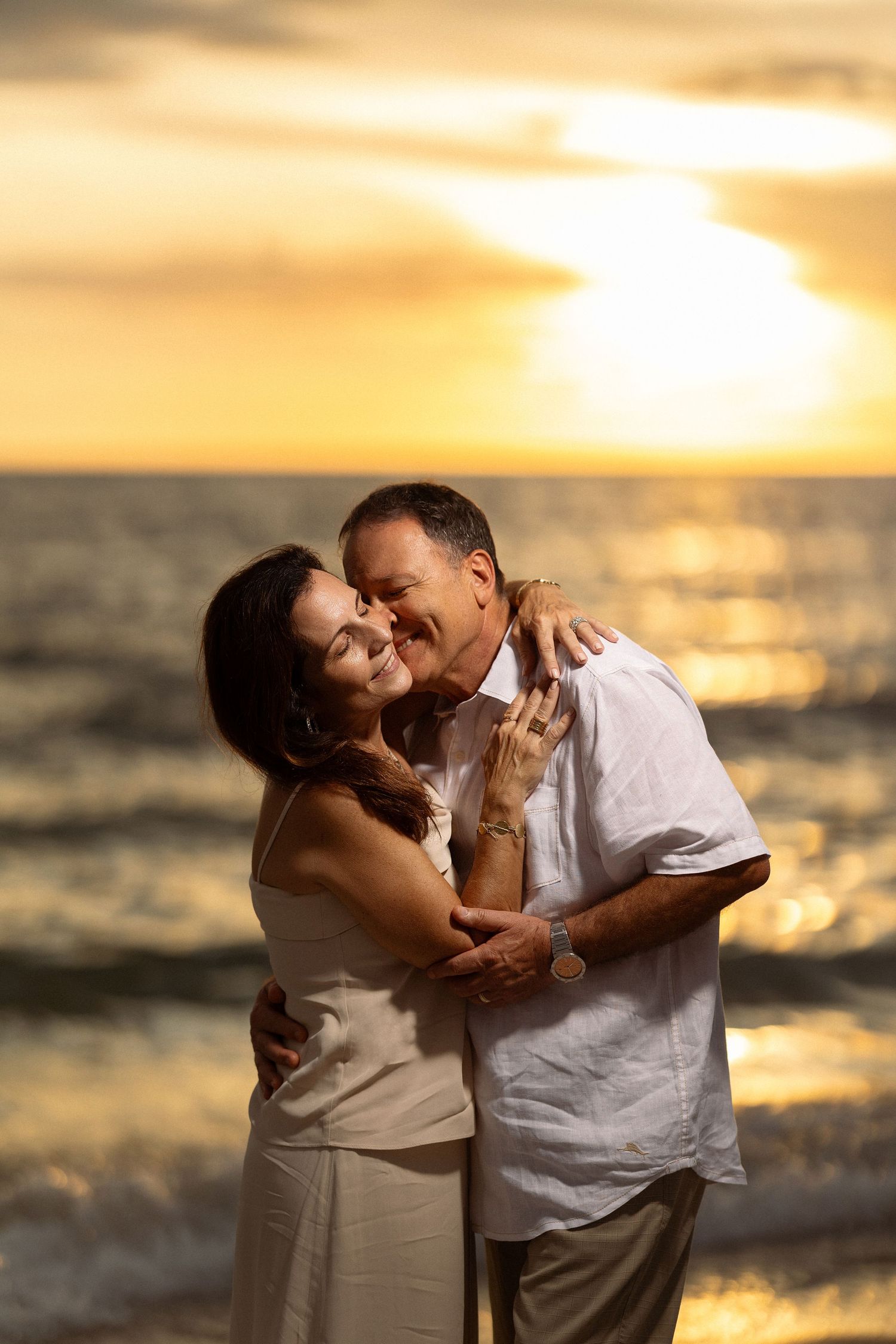 Couple embracing on beach at sunset with golden light reflecting off ocean waves.