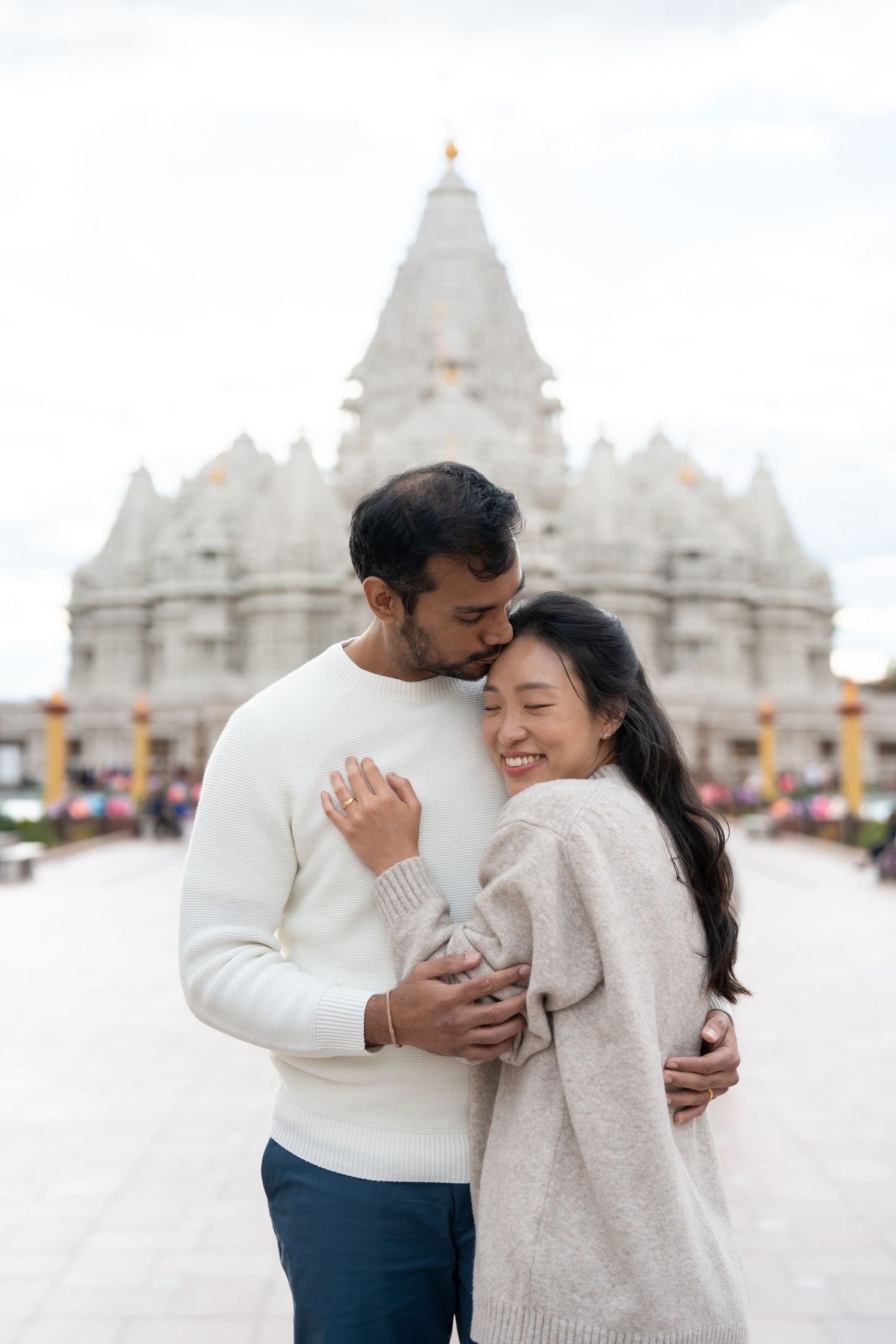 A romantic couple embraces and laughs together in front of white temple spires on a bright cloudy day.