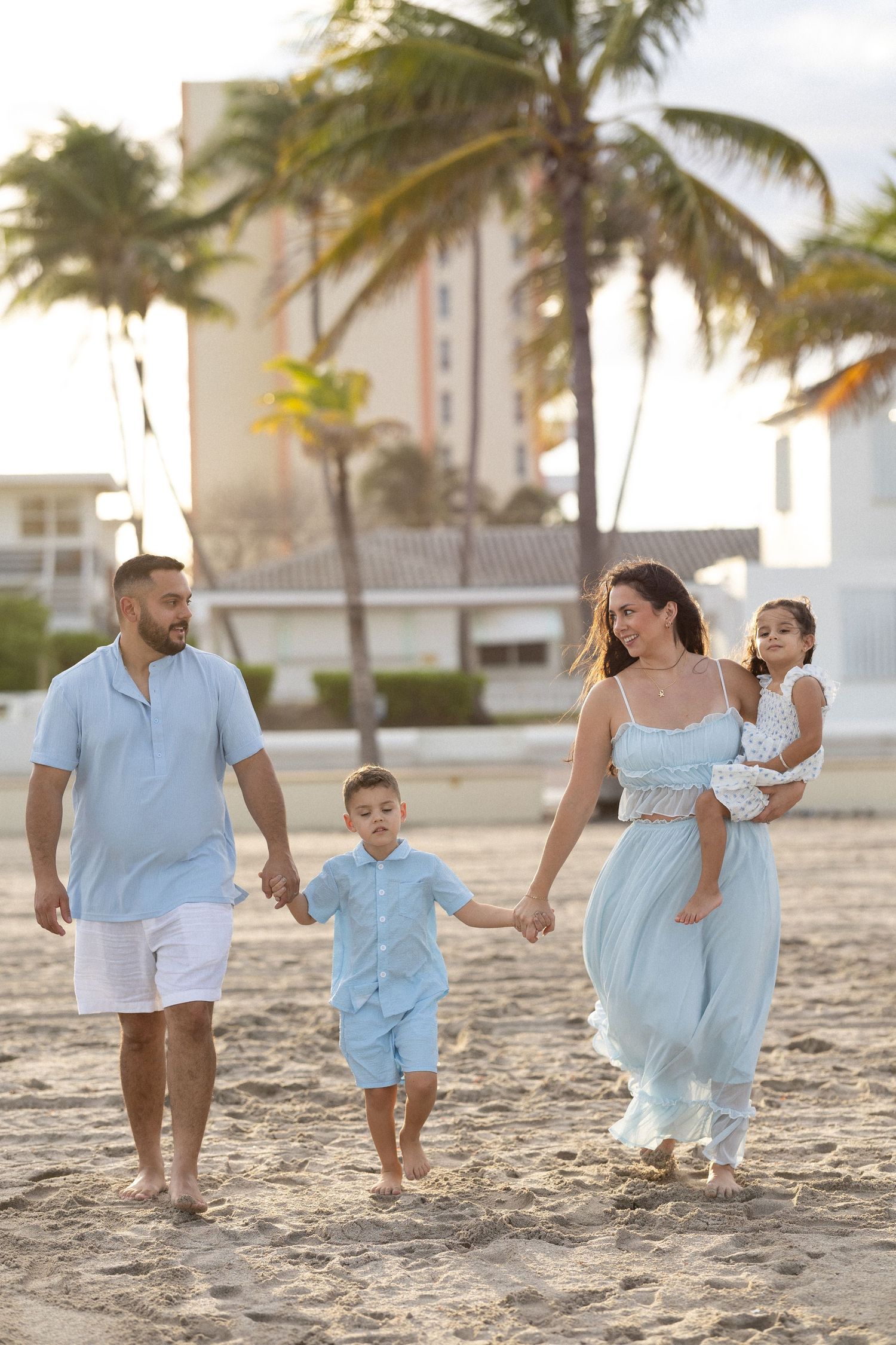 Family walking together on a beach at sunset with palm trees and buildings in the background.