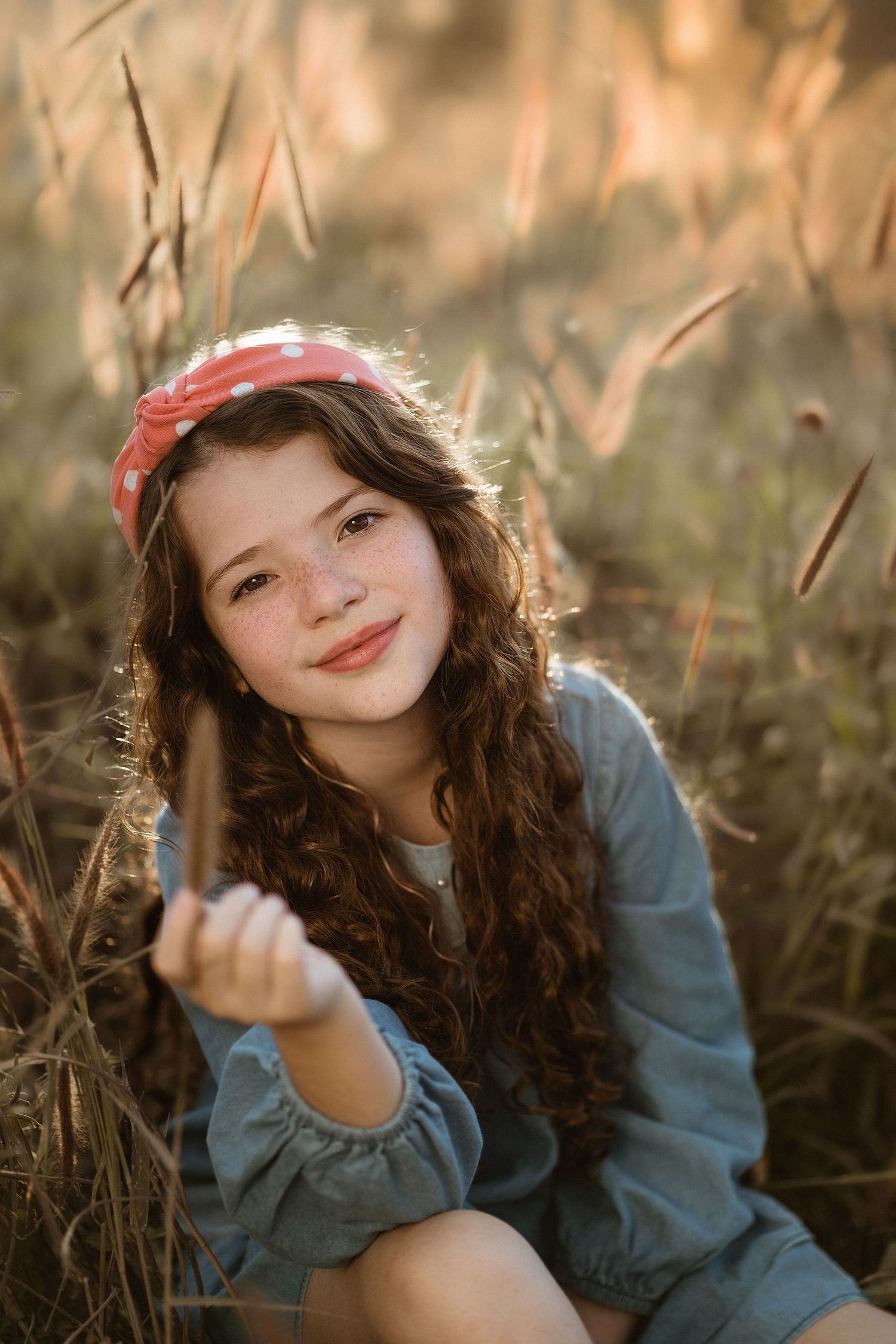 Young girl with curly hair and red headband posing playfully in tall grass at golden hour.