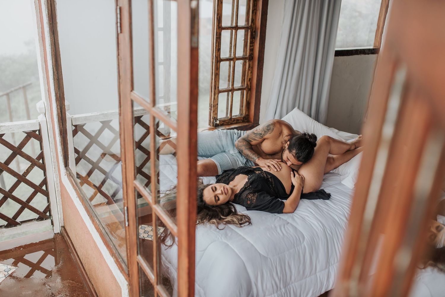 Romantic couple lying together on a bed with wooden lattice window frames in the background.