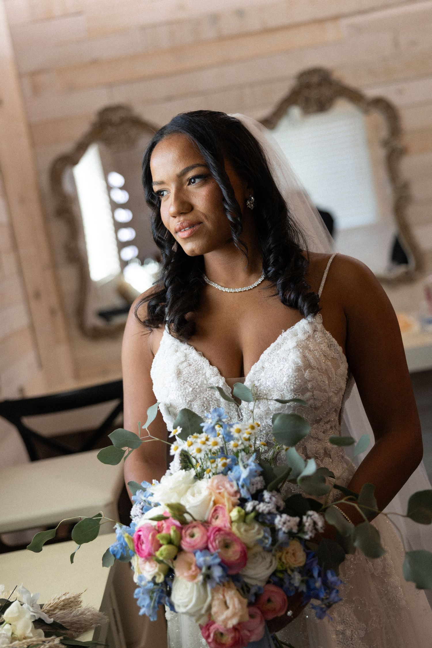 A bride in a white lace wedding dress holds a vibrant bouquet of pink, blue and white flowers in an elegant indoor setting.