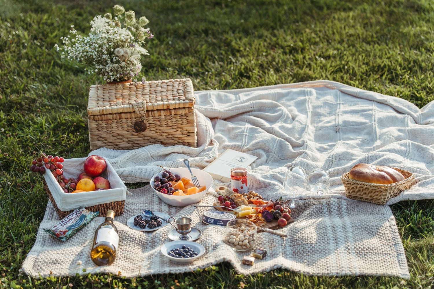 Picnic blanket laid out on grass with fresh fruits, bread and wicker basket.