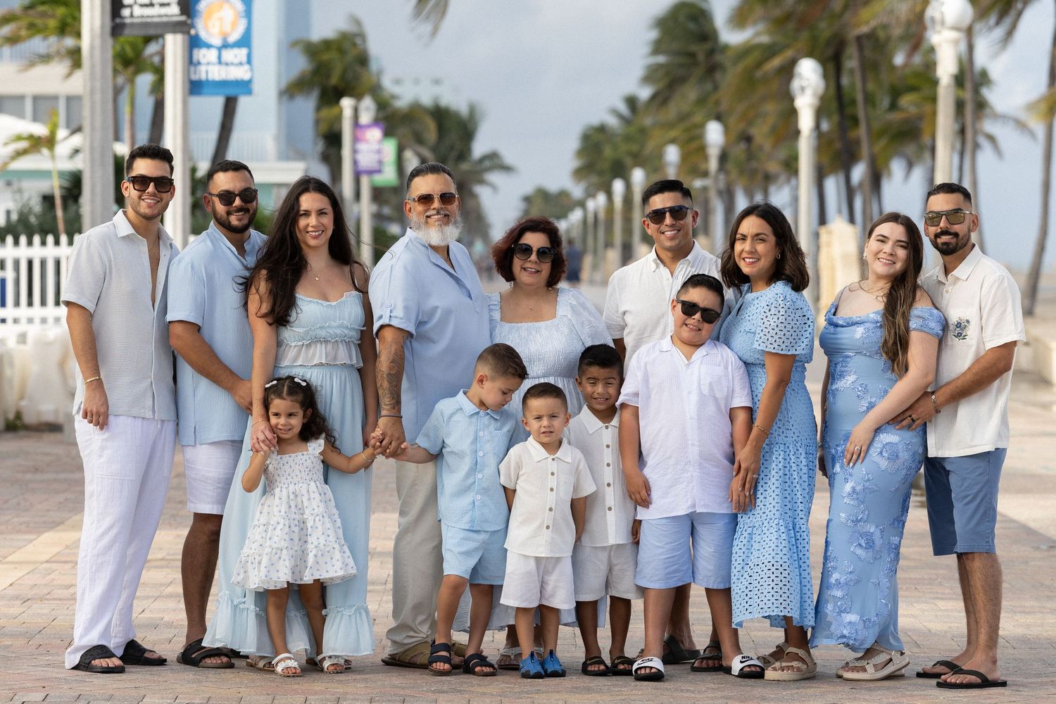 A large family group wearing coordinated blue and white outfits poses together on a palm-lined beach walkway.