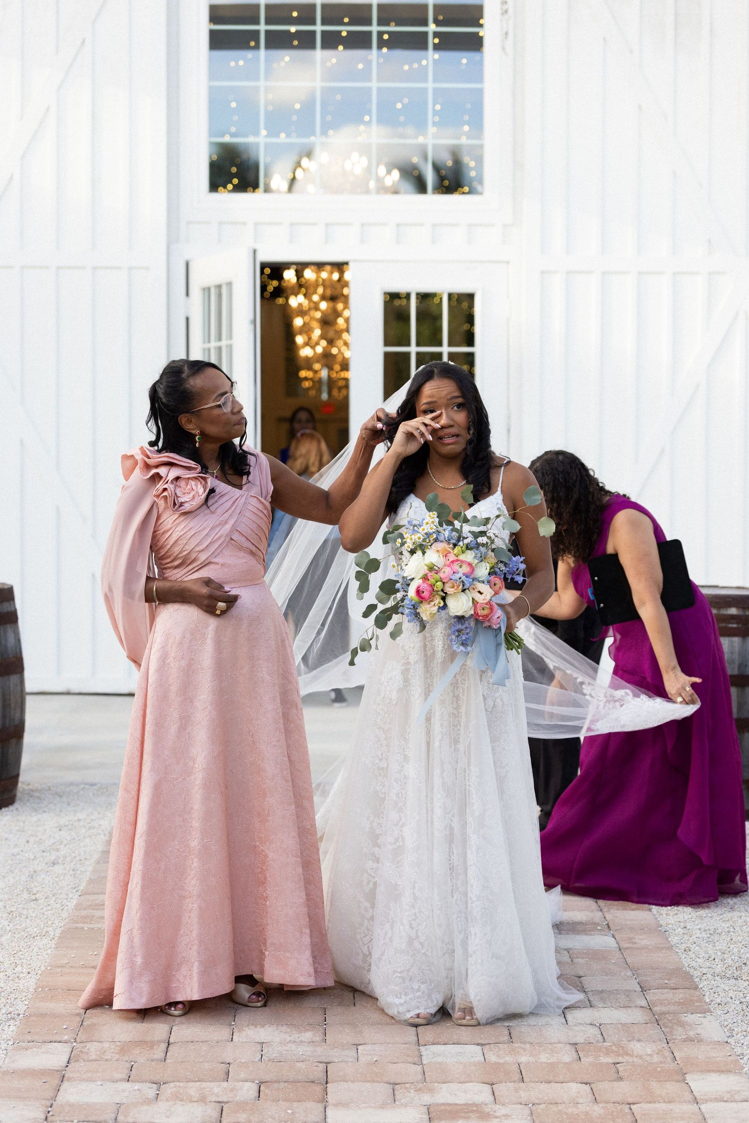 Bridesmaids in pink and purple gowns share an emotional moment with bride in white dress holding bouquet outside venue.